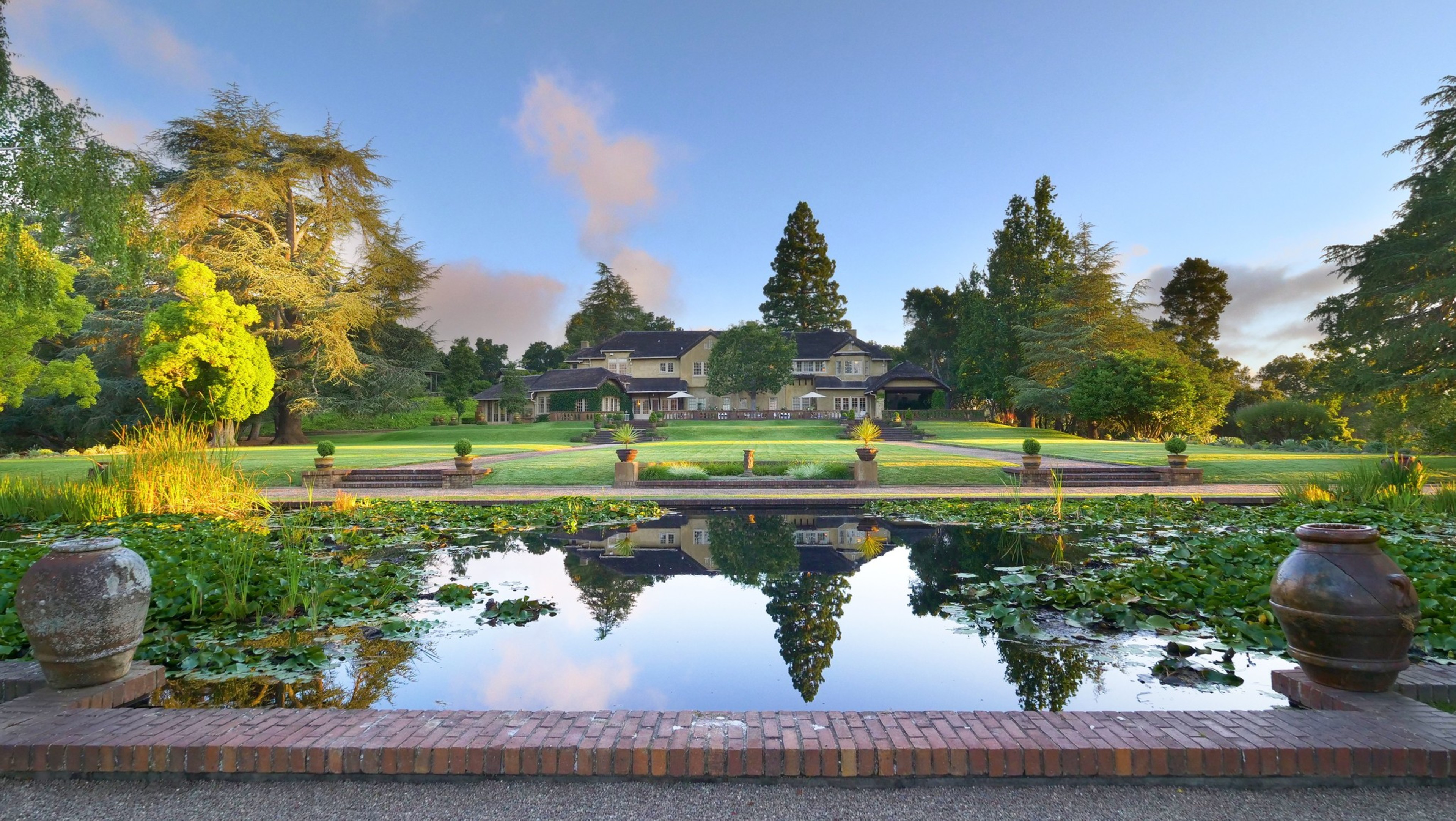 A large house sits behind a rectangular pond with lily pads, surrounded by manicured lawns, trees, and two large urns on either side of the pond.