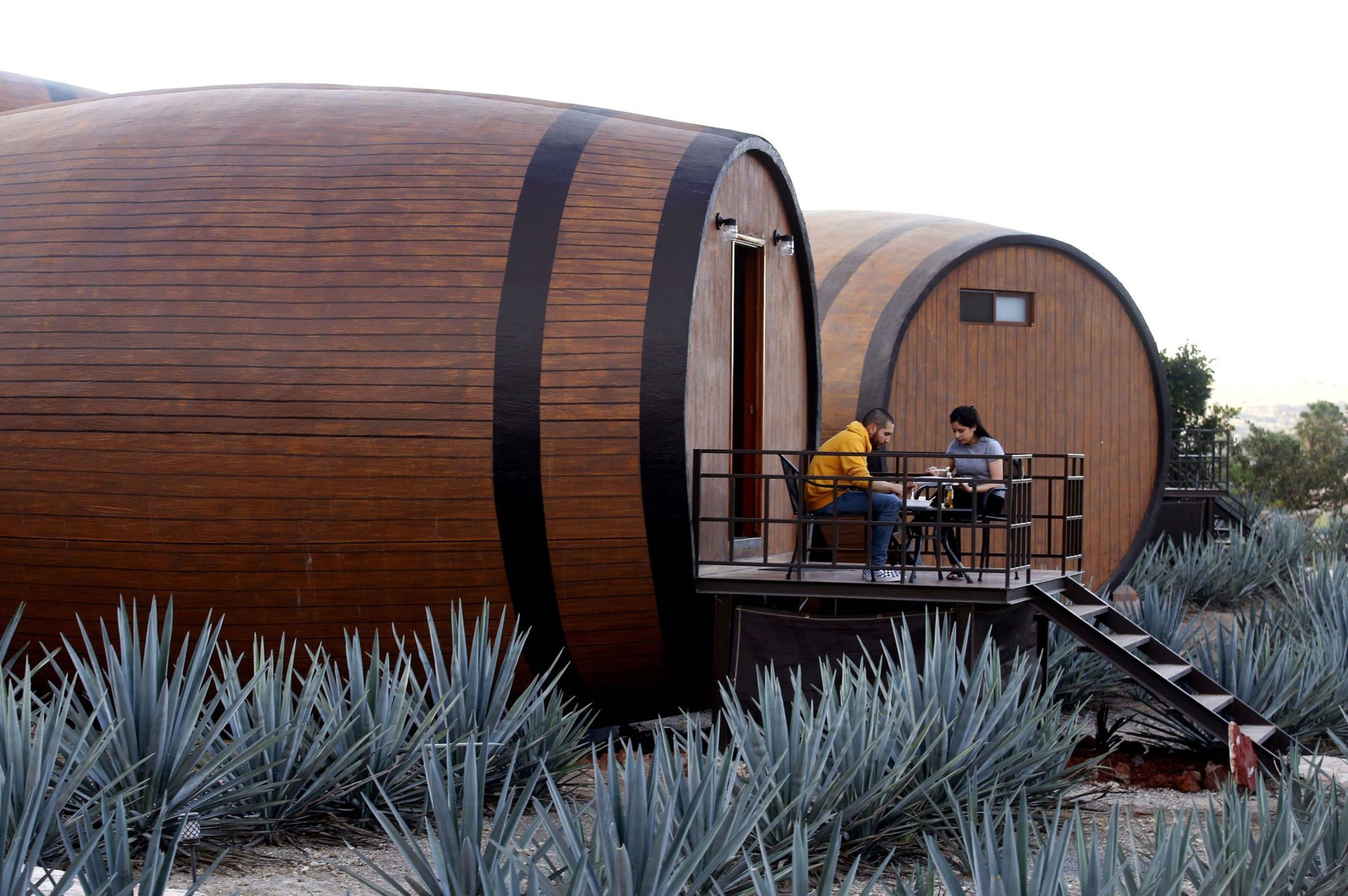 Two people sit at a table on a small porch attached to a building shaped like a giant barrel. Agave plants surround the area, creating a unique ambiance.