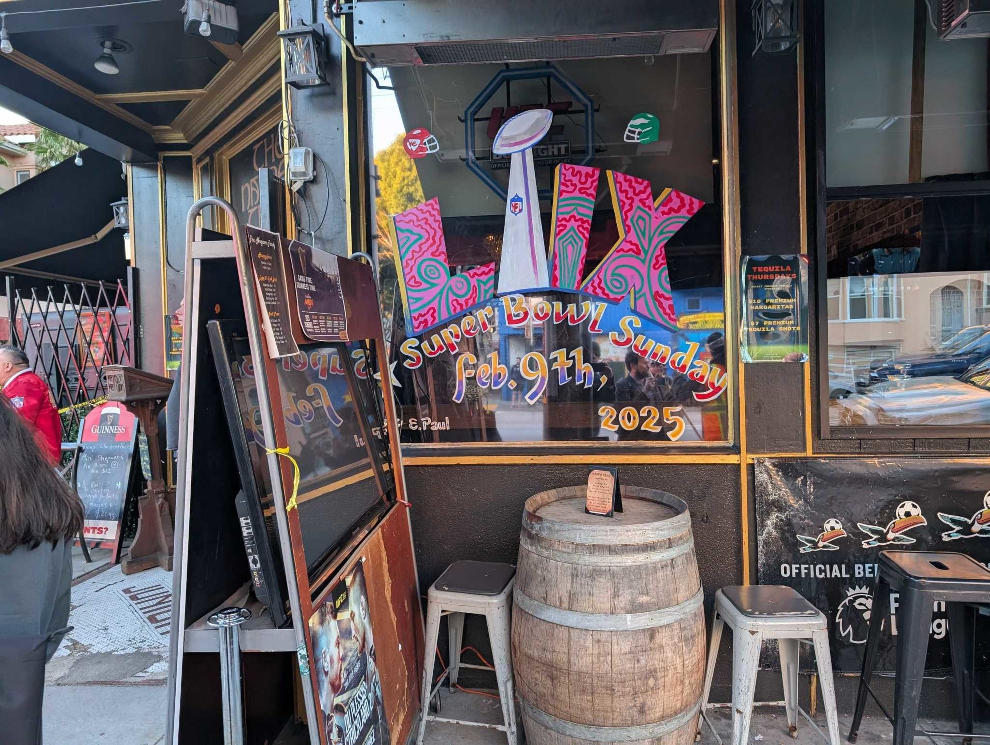 A storefront displays vibrant art with &quot;Super Bowl Sunday, Feb. 9th, 2025&quot; alongside a football trophy graphic, a barrel table, and stools outside.
