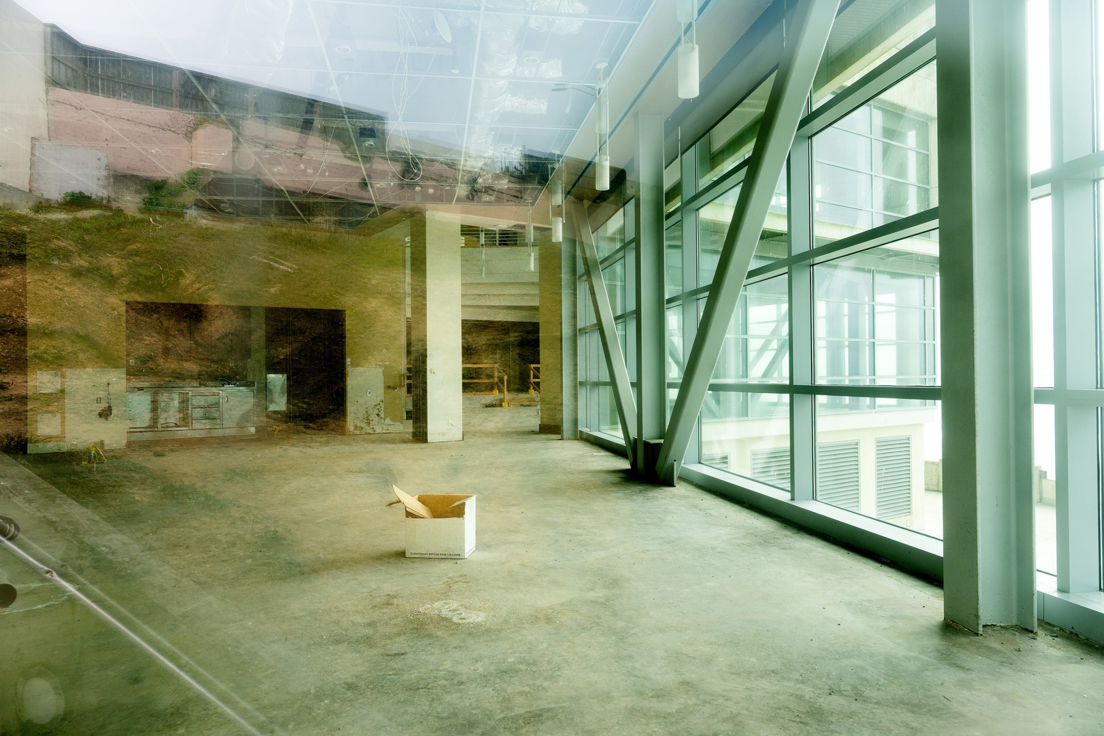 An empty, unfinished room with large floor-to-ceiling windows on one side and a single open cardboard box on the concrete floor.