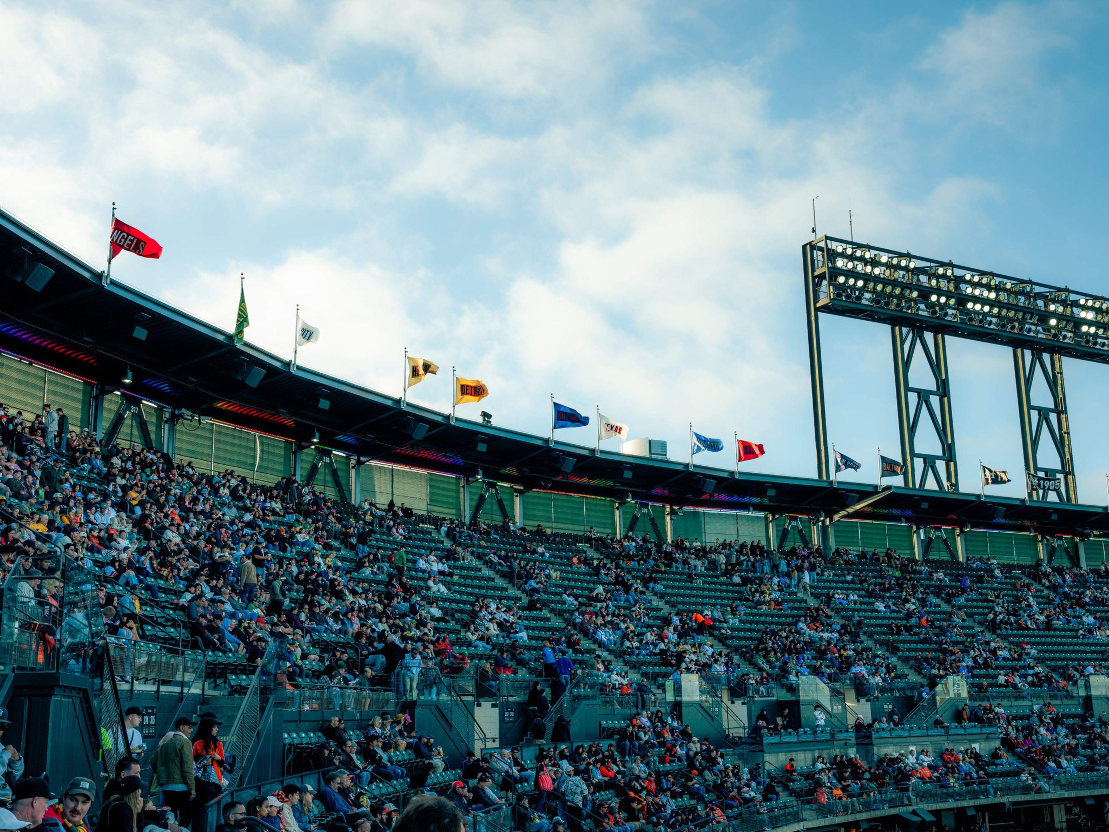A partially filled baseball stadium stands under a cloudy blue sky with several team flags flying above the upper seating rows.