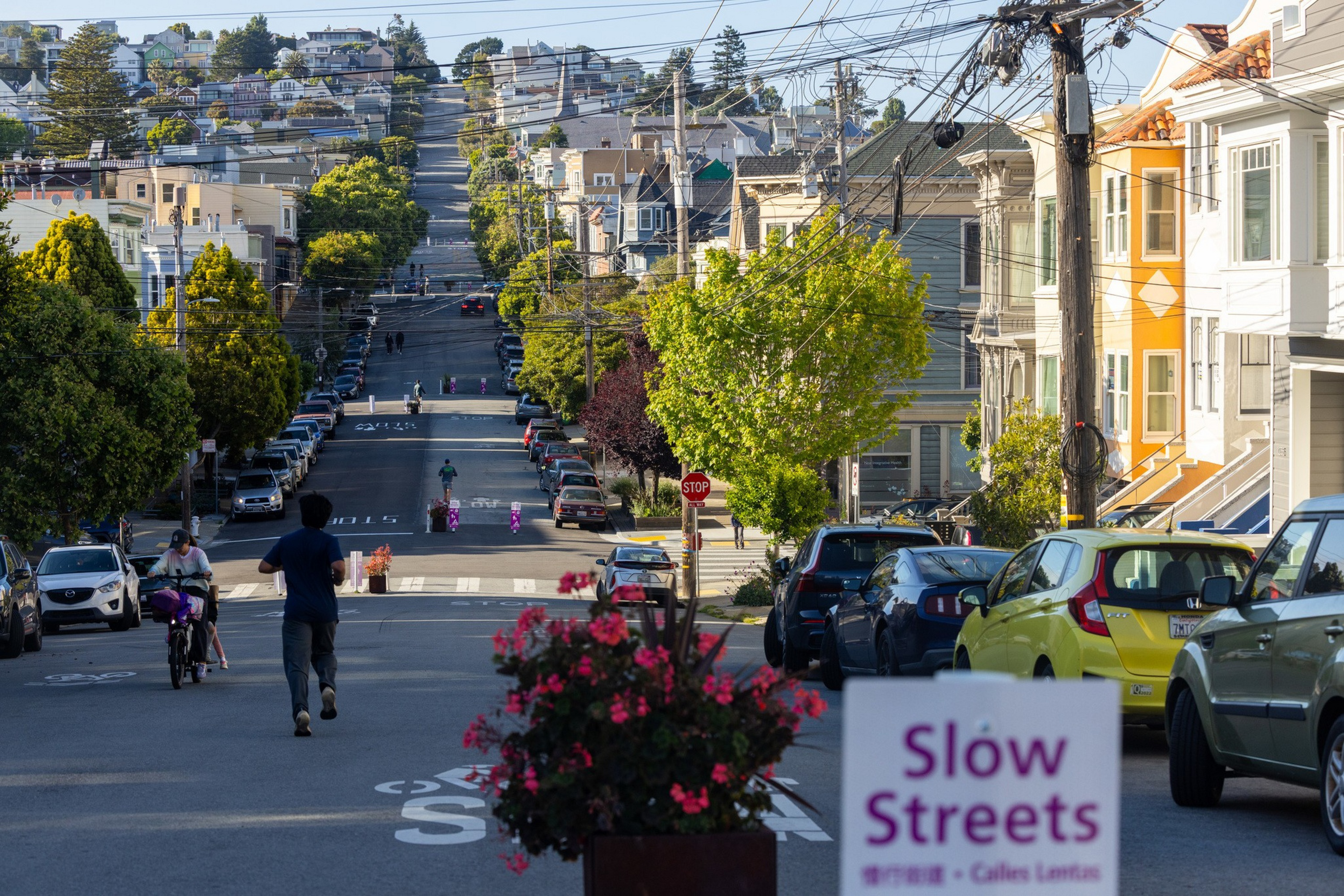 A street with trees, parked cars, and houses on both sides has people walking and biking, with a “Slow Streets” sign and flowers in the foreground.