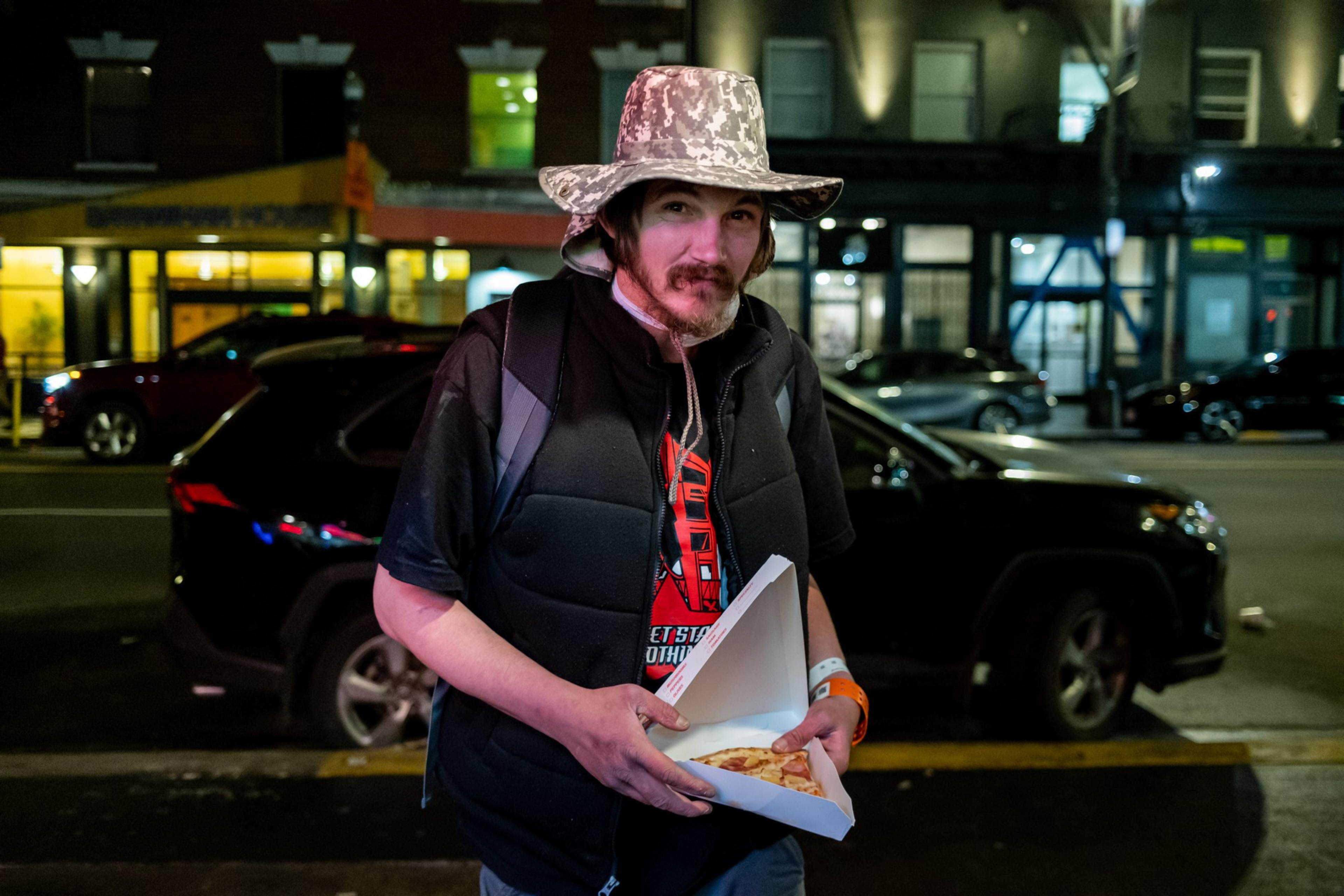 A person in a camo hat and black vest holds a pizza slice box on a nighttime city street with cars and lit buildings in the background.