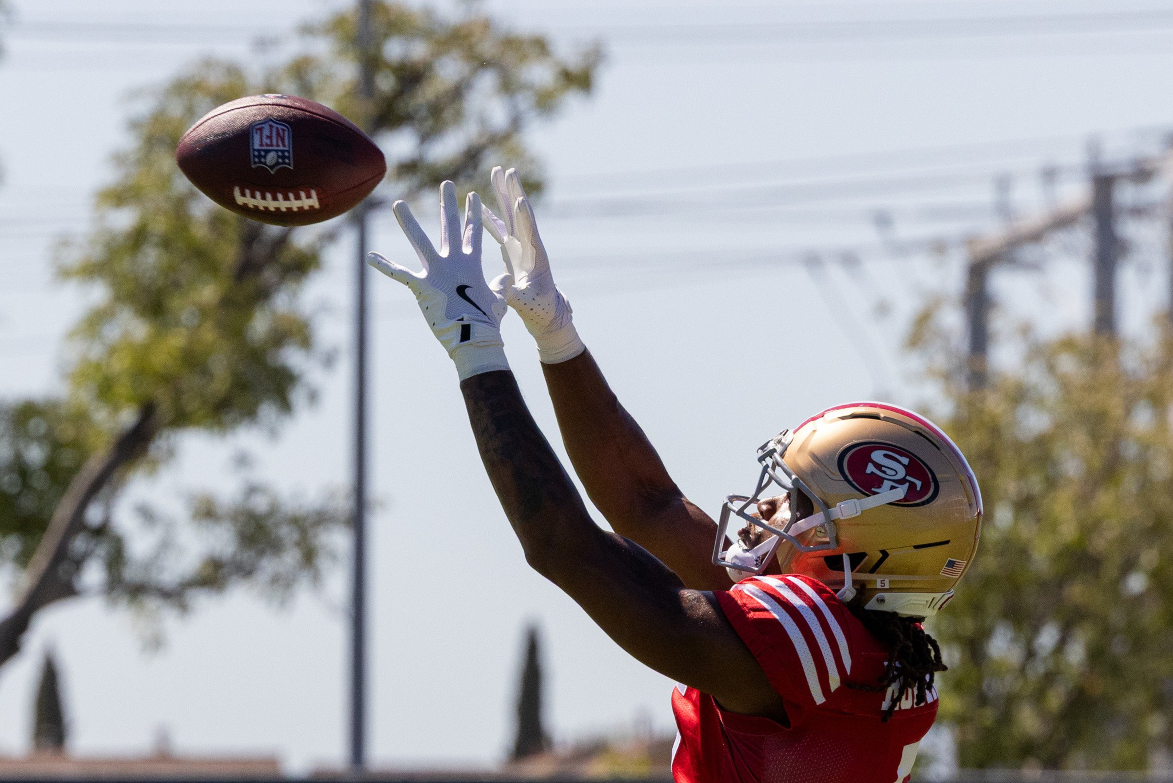 A football player in a red San Francisco 49ers uniform and gold helmet reaches up with both hands to catch an NFL football in midair.