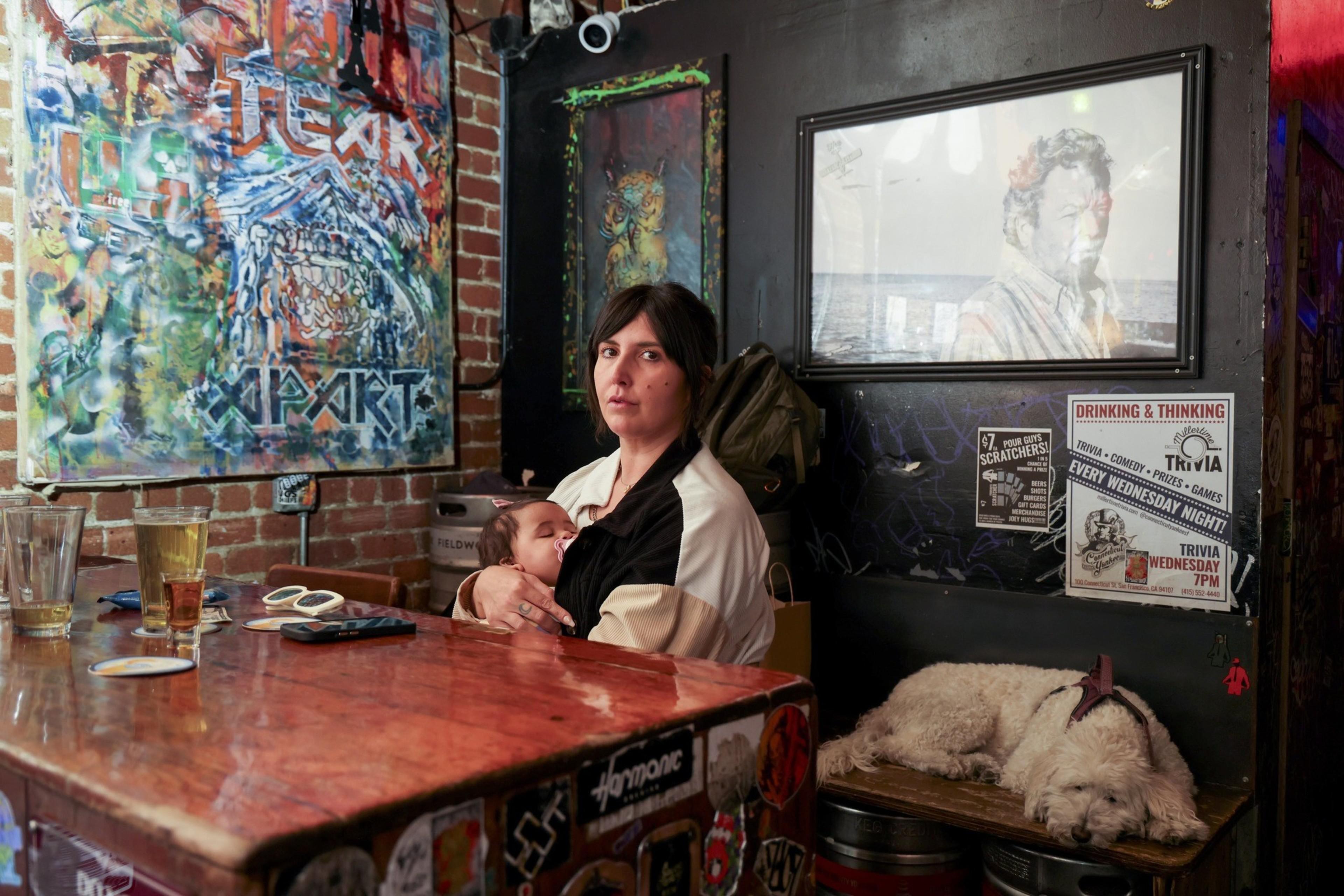 A woman holds a sleeping baby in a colorful bar with art on the brick walls. A fluffy dog sleeps on a bench beneath posters advertising trivia night.