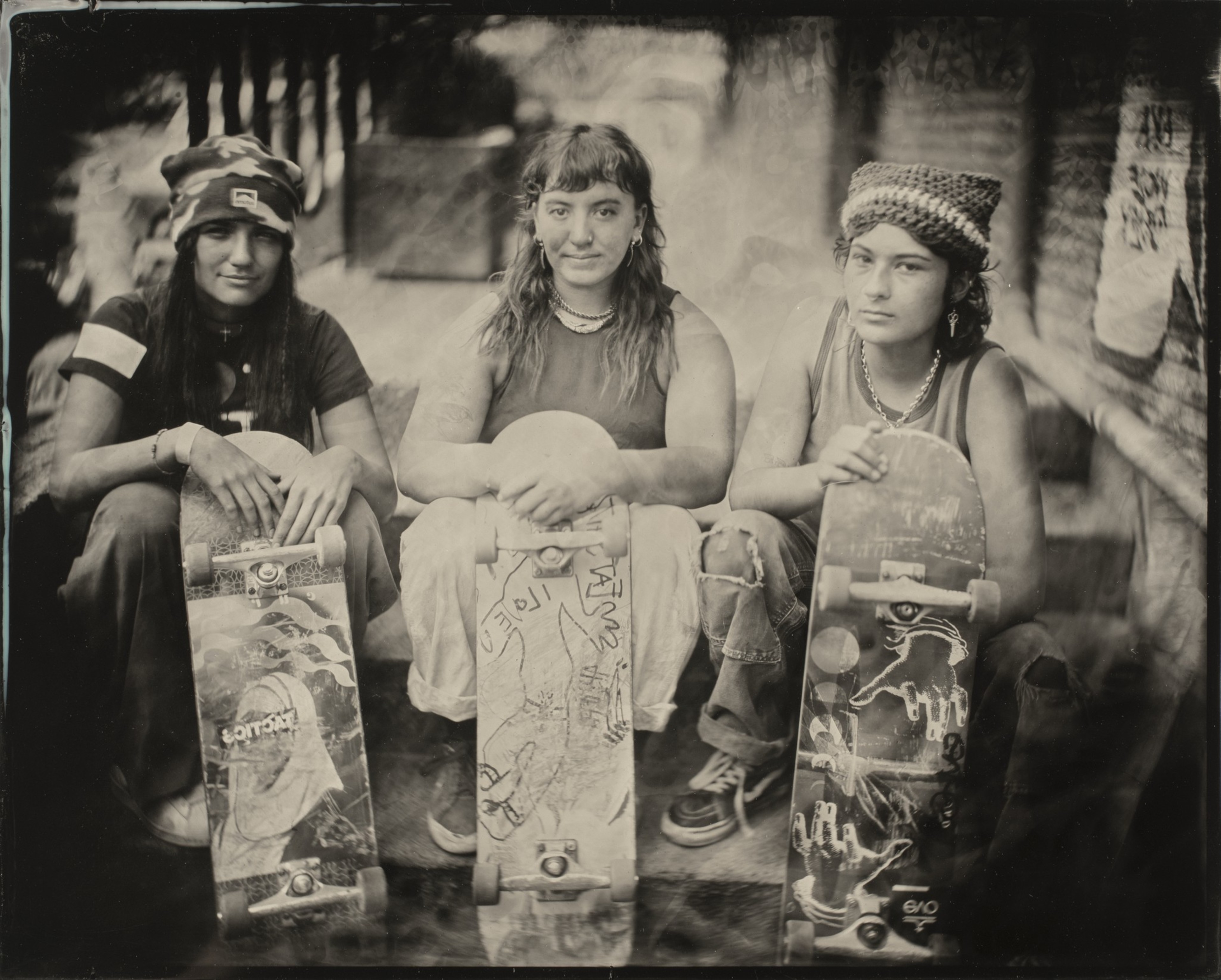 Three people sit side by side, each holding a skateboard vertically. They are dressed casually and look directly into the camera. The image is in black and white.