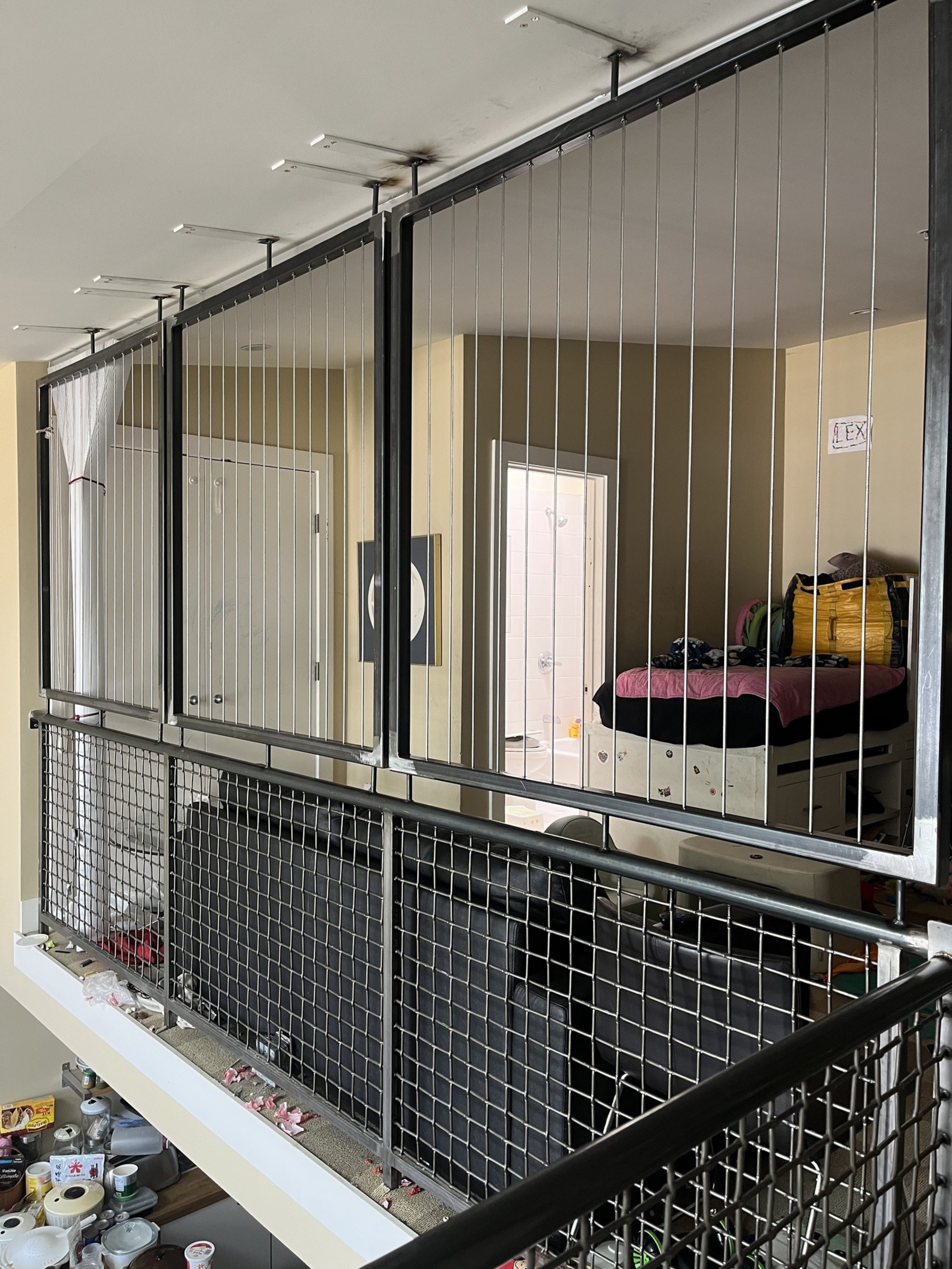 A loft-style room with a black wire railing, a bed with pink bedding and bags on it, a white door, and various items scattered on a lower level.