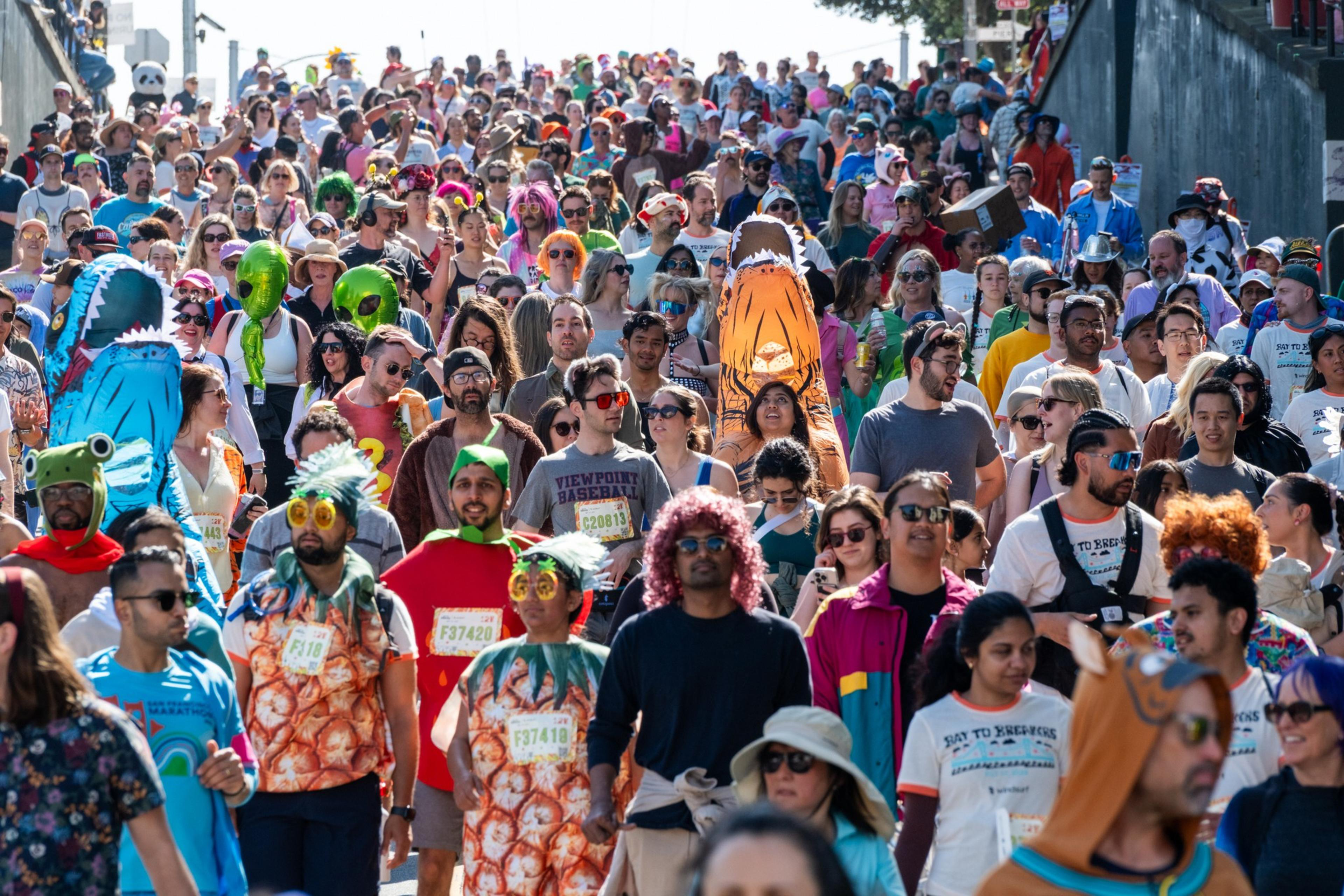 A vibrant, colorful crowd of people in costumes, including sharks and aliens, are walking outdoors in a festive, lively atmosphere.