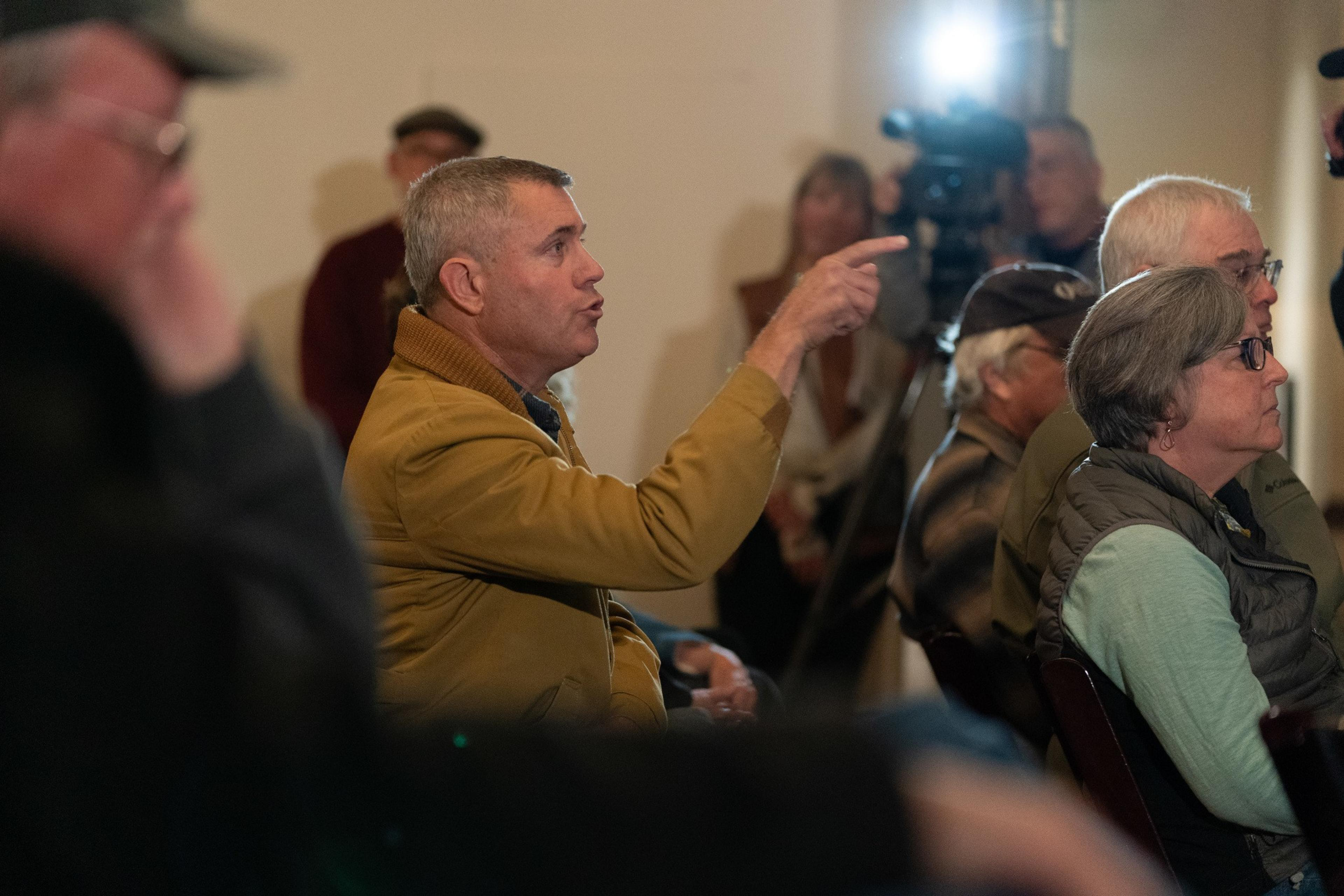 A person points from his chair in a tan jacket.