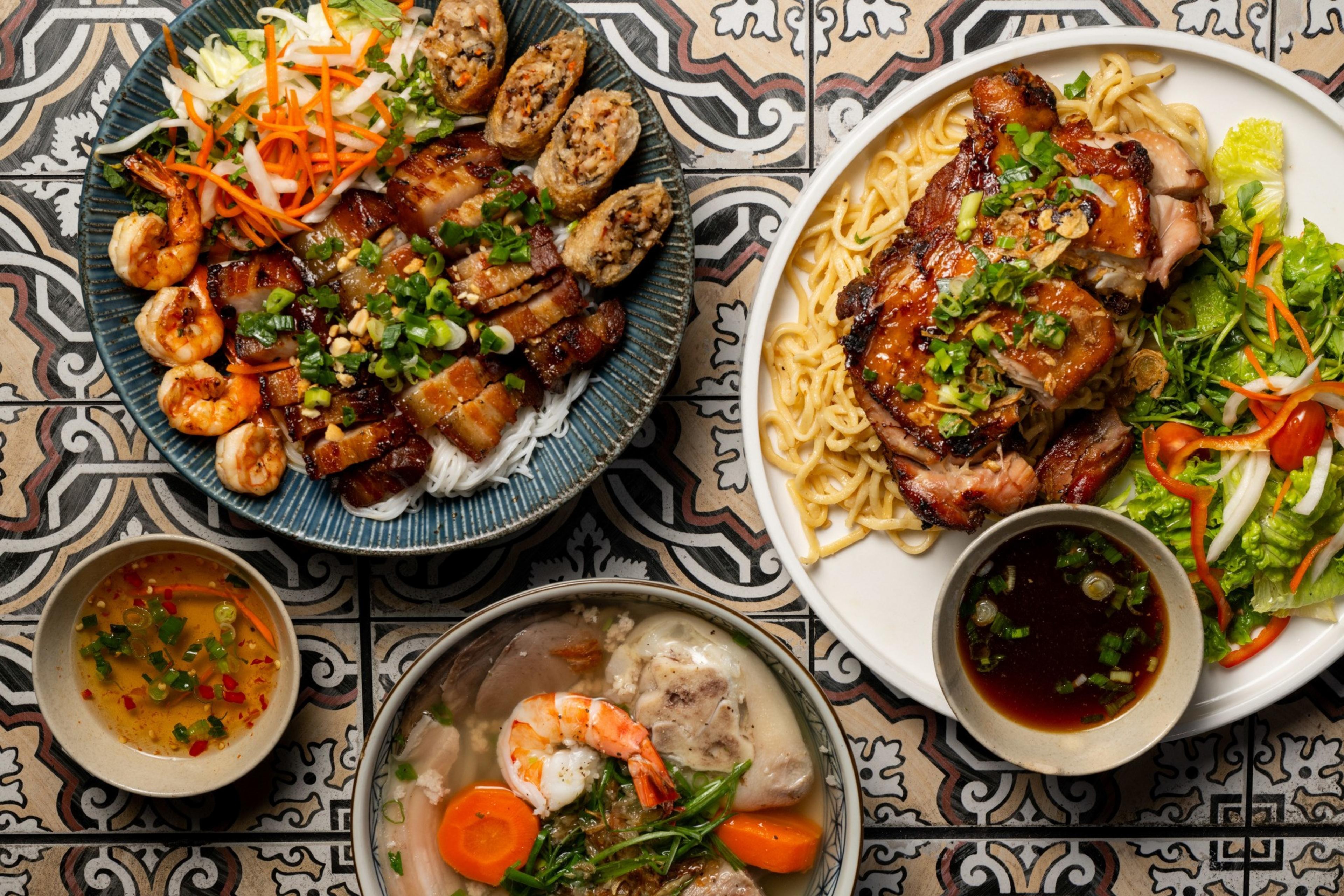 The image shows three plates of Vietnamese cuisine with noodles, grilled meats, shrimp, fresh vegetables, and spring rolls; accompanied by two small bowls of dipping sauces.