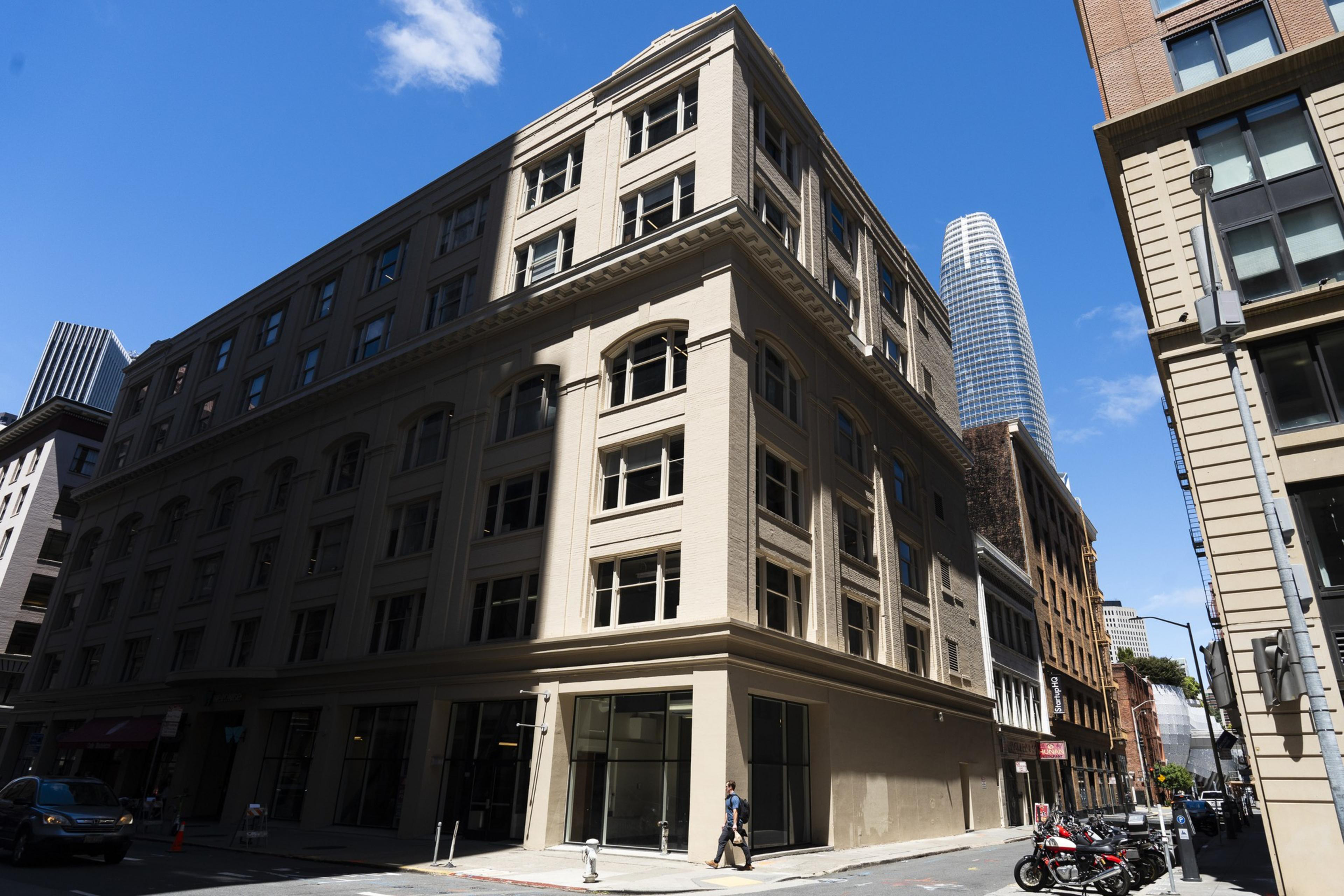 A beige, multi-story building stands on a sunny street corner with a blue skyscraper visible in the background. A person walks by, and motorcycles are parked nearby.