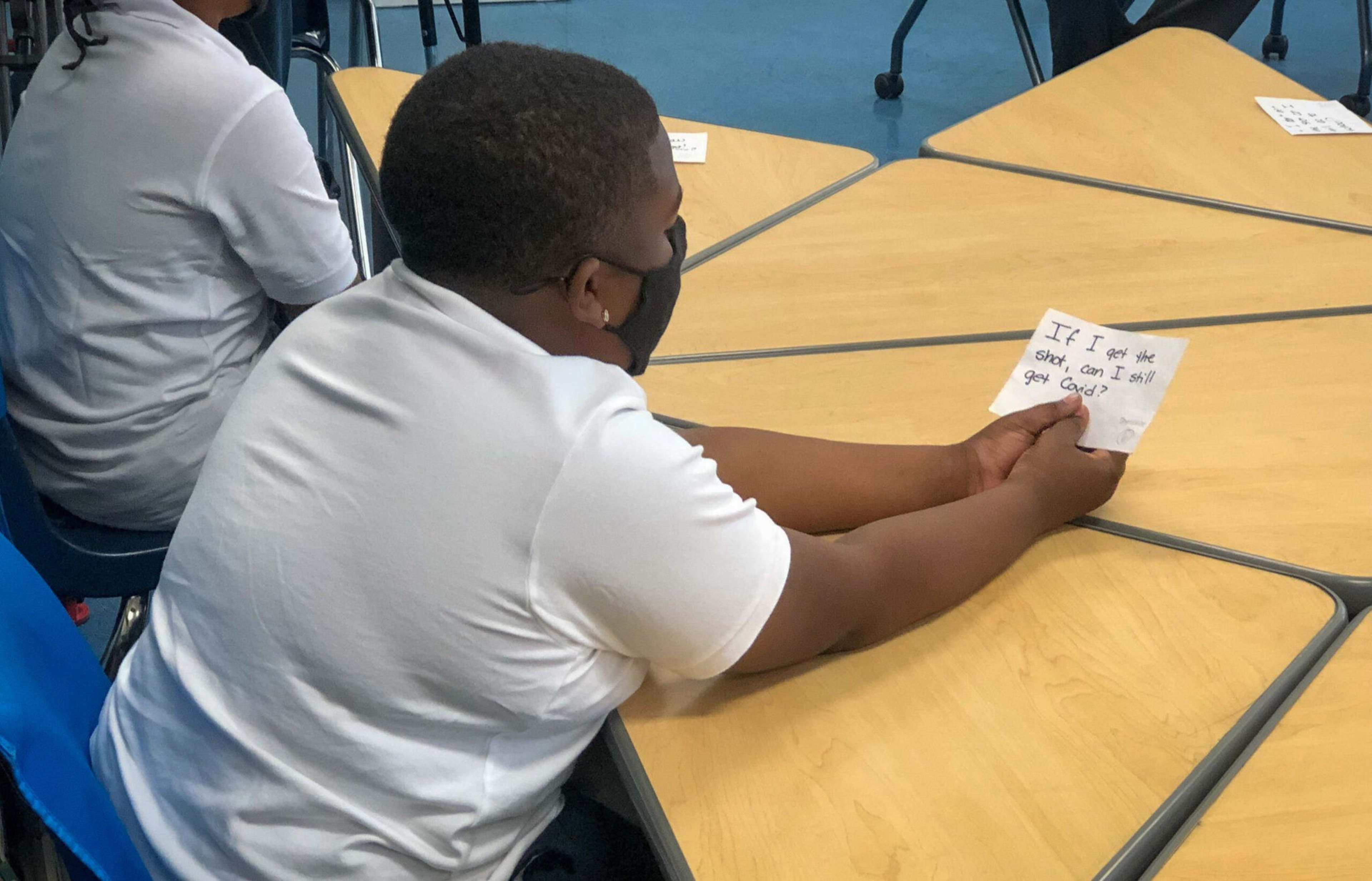 A boy wearing a mask sits at a table holding a note that reads, "If I get the shot, can I still get Covid?" in a classroom setting.