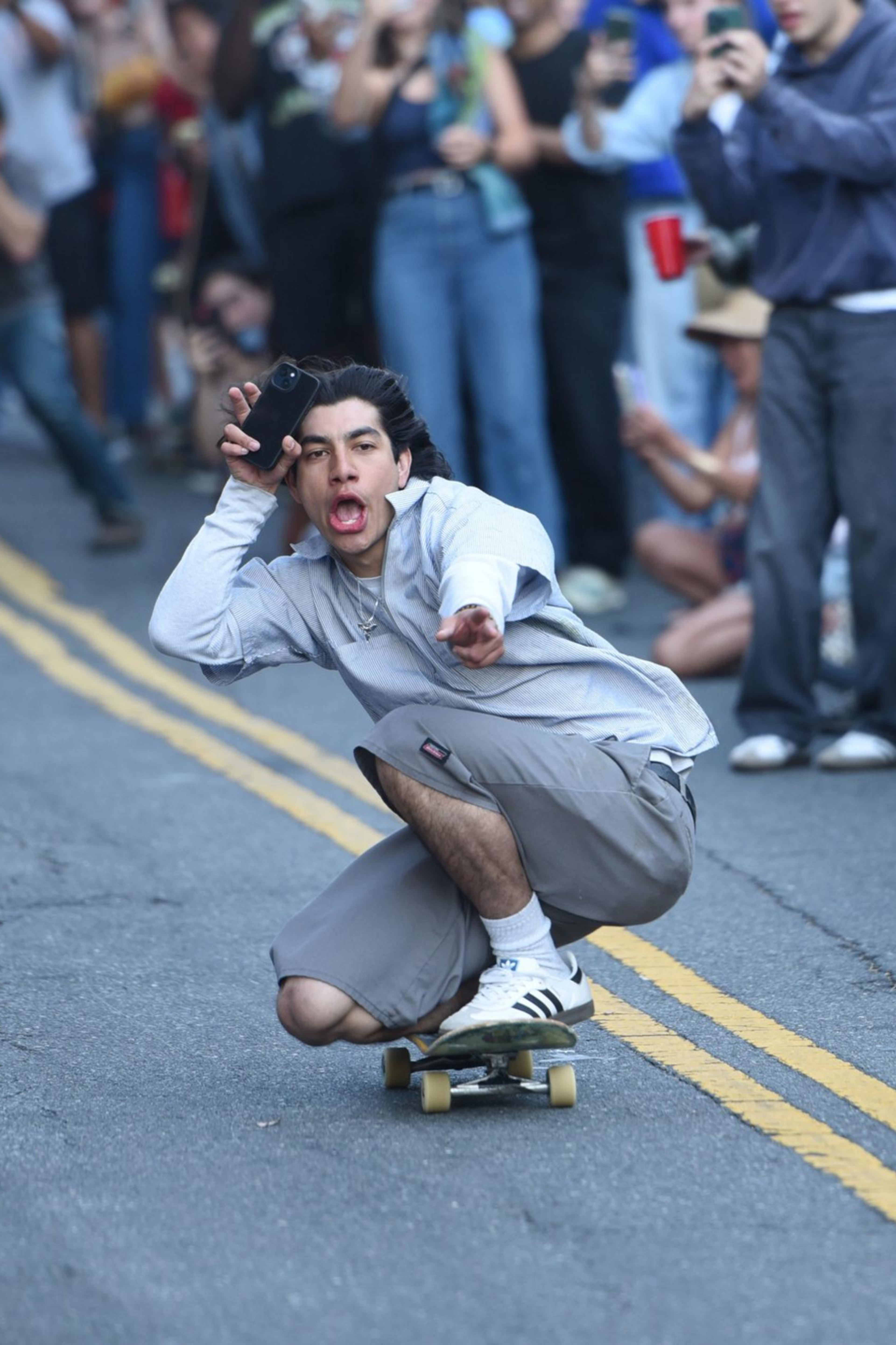 A man skateboards down a street, crouching low with a playful expression, while holding a phone up. A crowd of onlookers, many taking photos, lines both sides of the street.