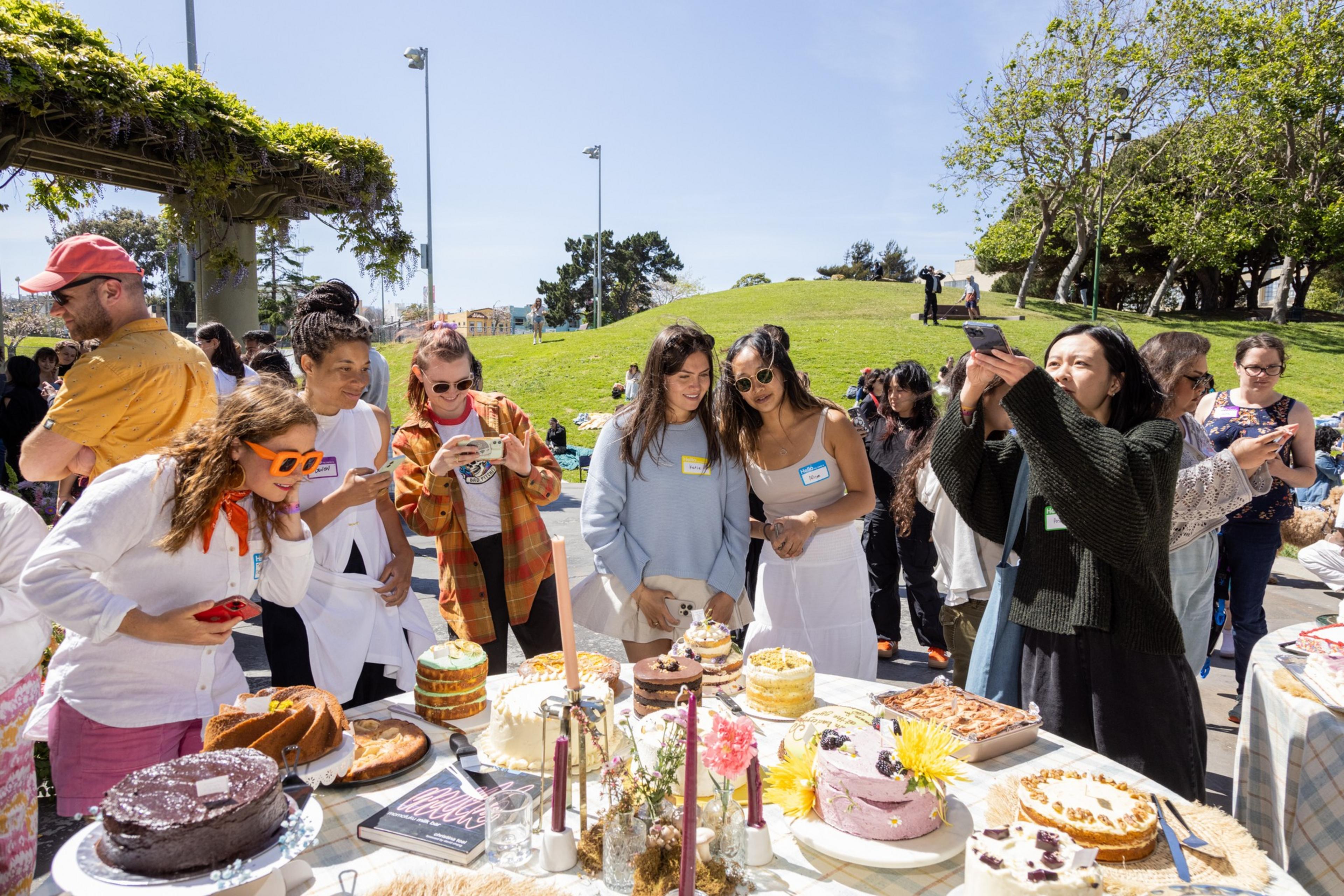 Outdoor cake contest with people inspecting and photographing various cakes on tables. Sunny park setting.