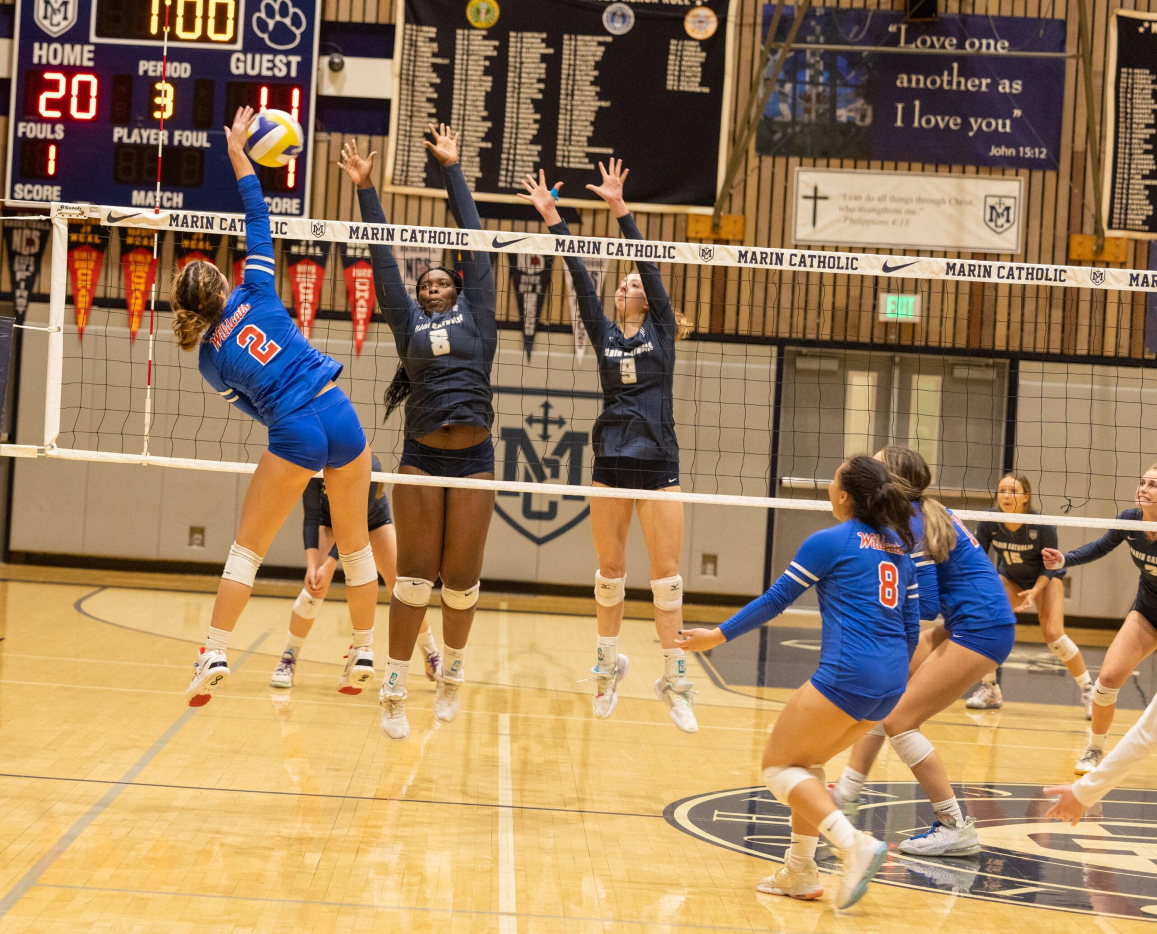 A volleyball player in blue jumps to spike the ball while two players in black jump to block at the net during a match with a 20-11 score in the third period.