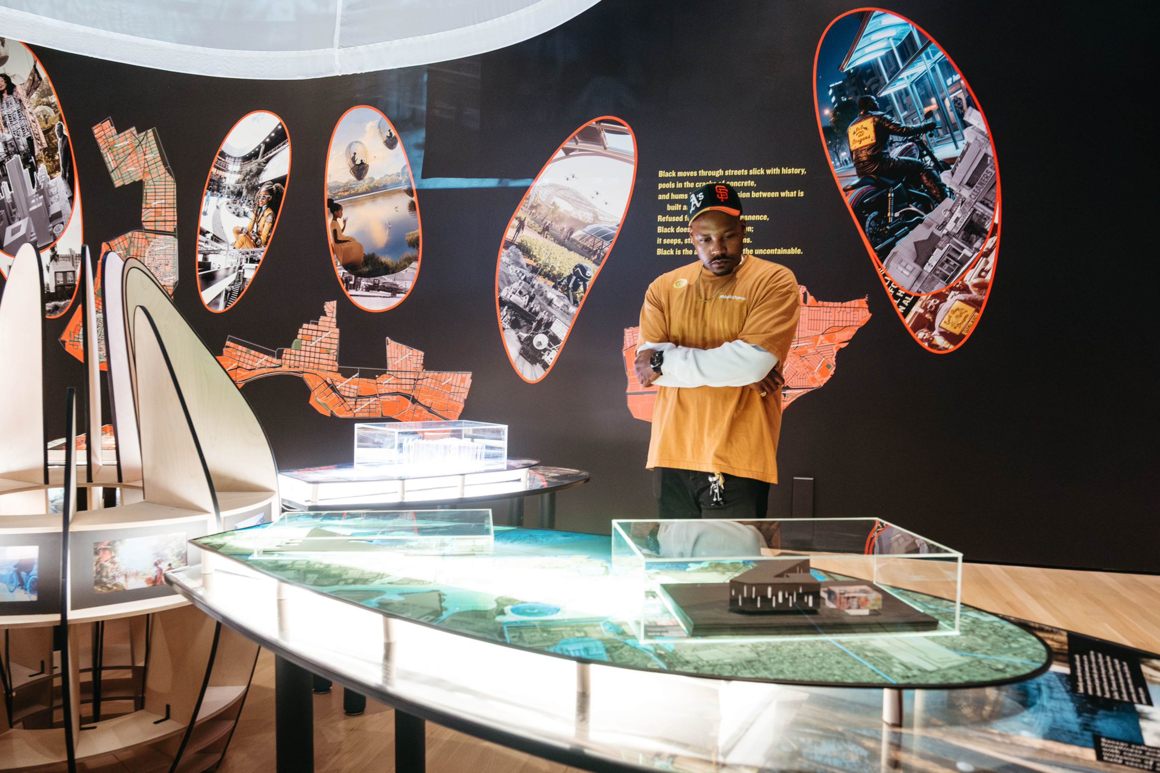 A man in an orange shirt stands with arms crossed in a modern exhibit featuring illuminated architectural models and colorful, oval-shaped wall displays.