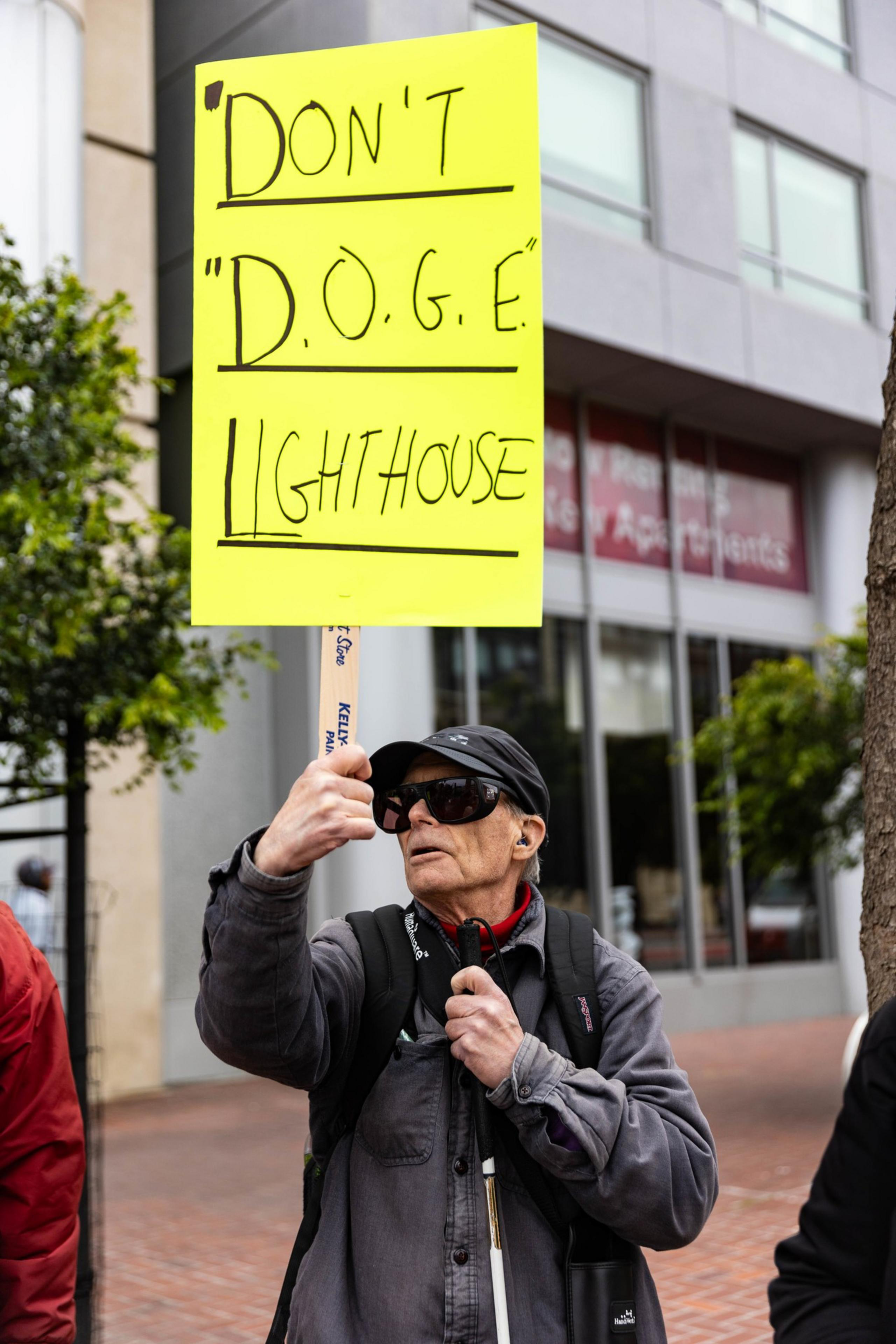A person in dark glasses holds a yellow sign saying, &quot;Don't 'D.O.G.E' Lighthouse,&quot; standing outdoors with a building and trees in the background.