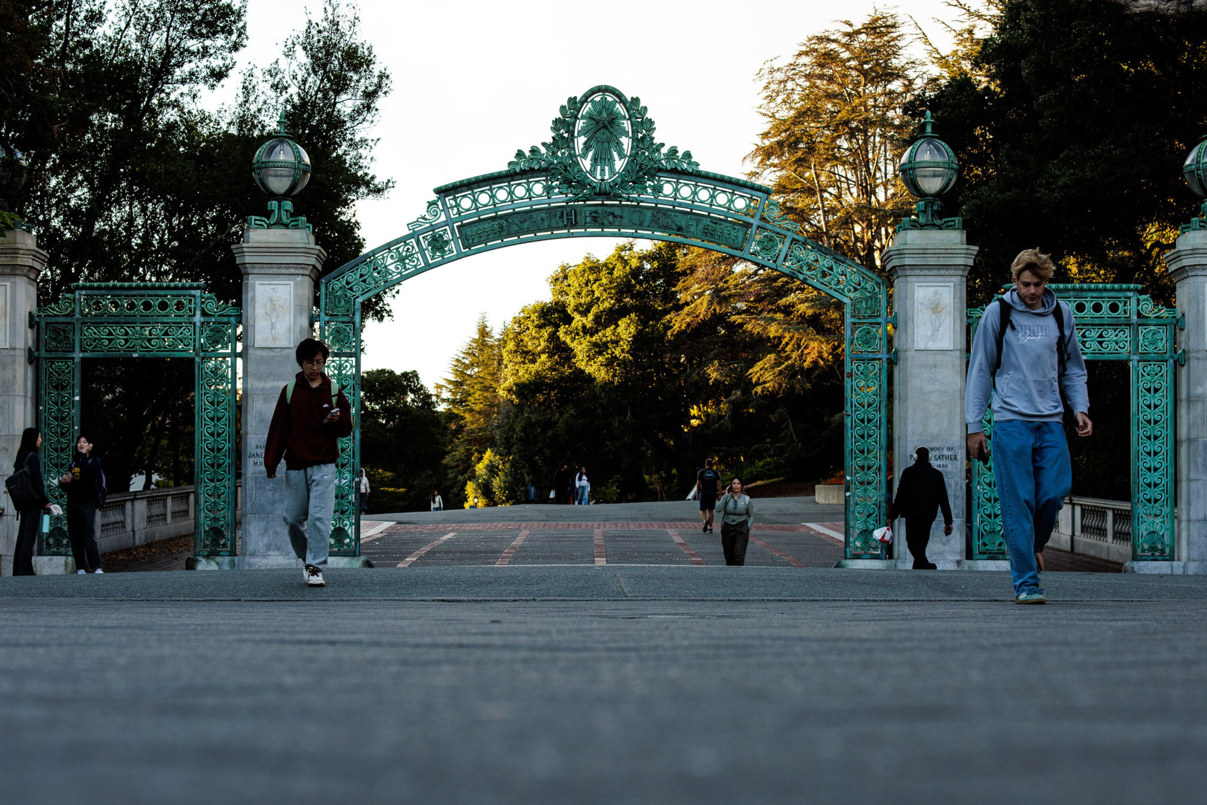 A decorative green iron archway stands over a paved path with people walking beneath it, surrounded by trees in the background.