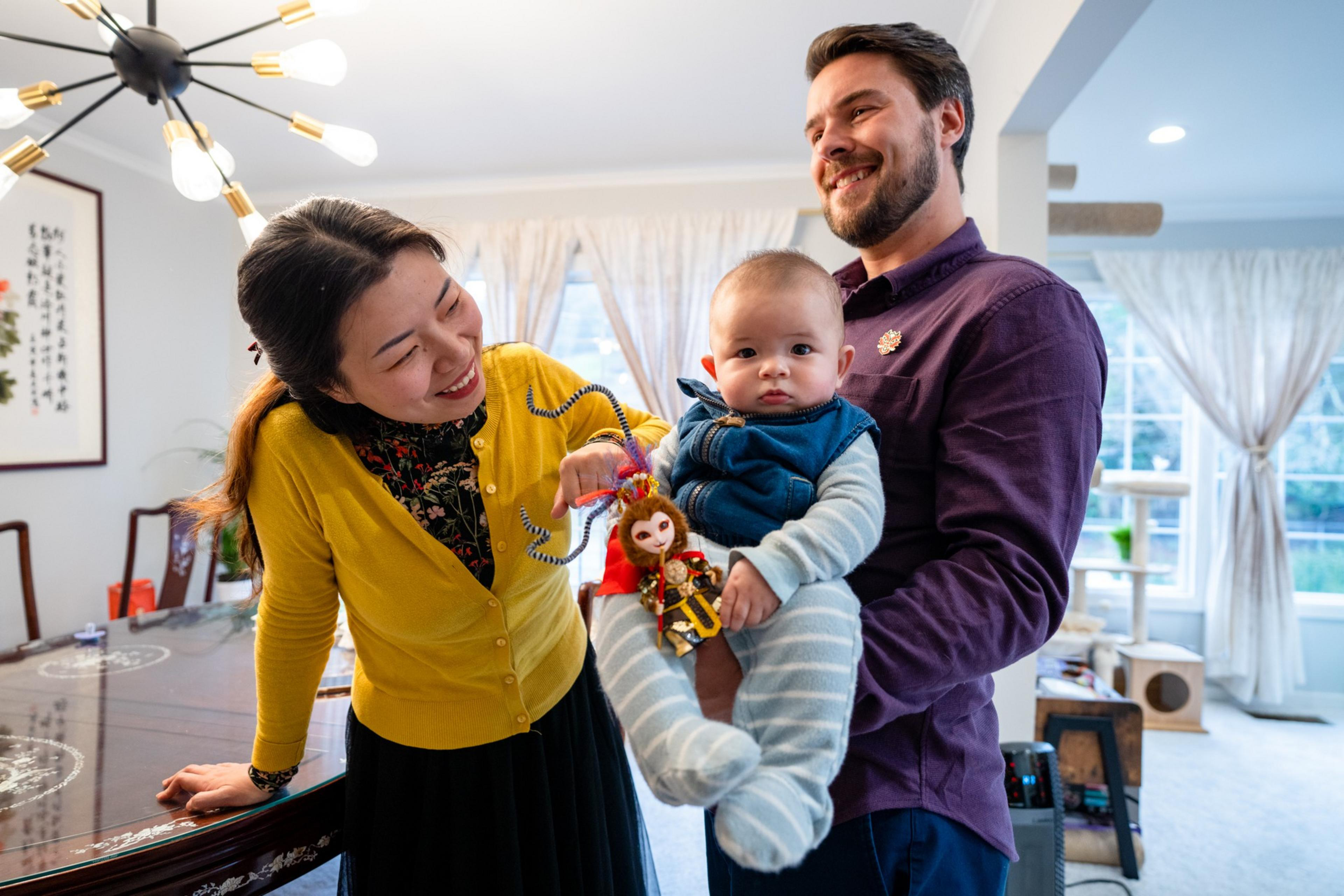 A smiling man holds a baby dressed in blue, while a woman in a yellow cardigan plays with a monkey doll watchfully near a dining table.