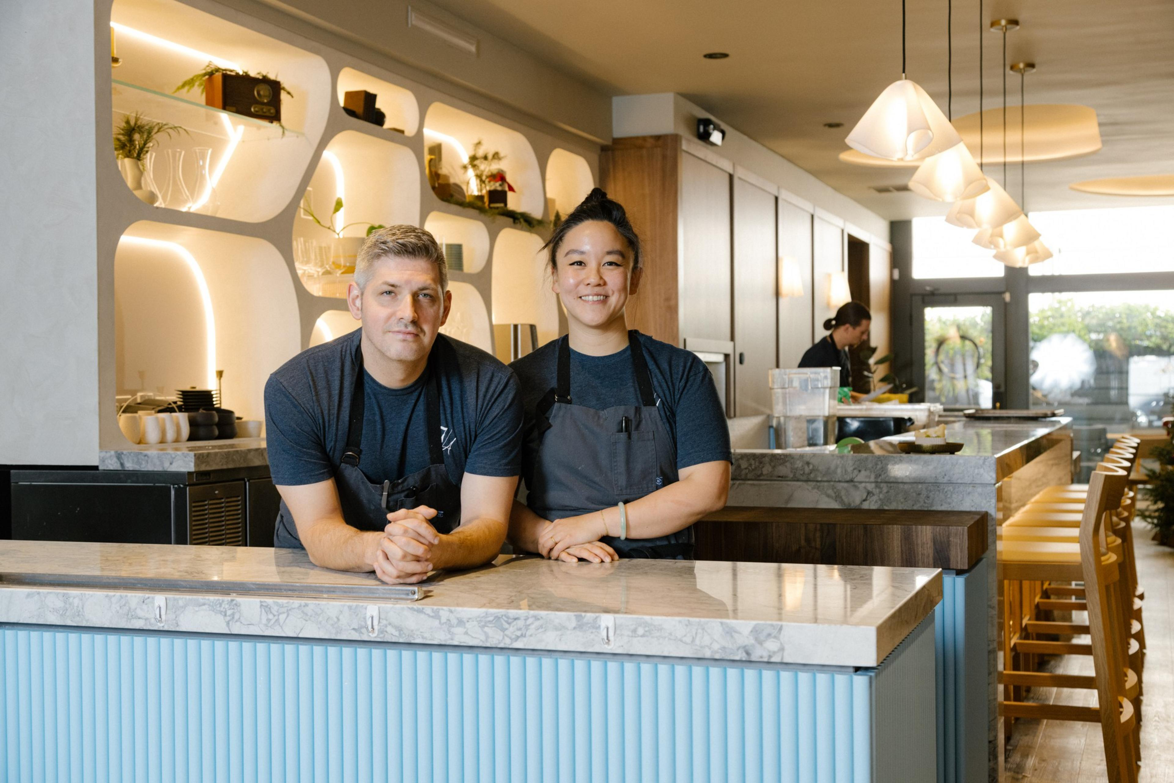 Two chefs smile from behind a cafe counter, with a modern, well-lit interior and decorative shelving in the background.