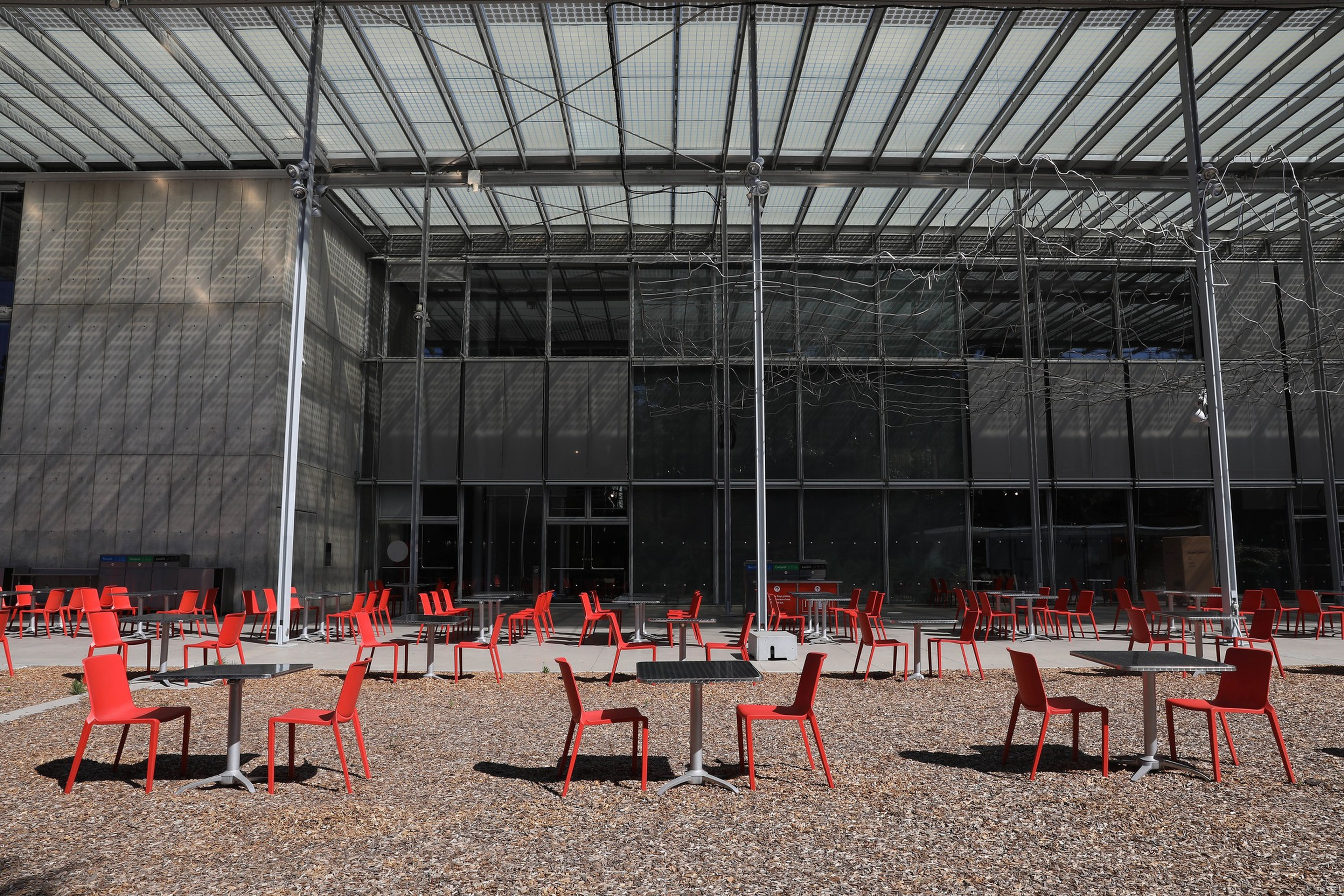 An outdoor seating area with multiple red chairs and round tables is set on a gravel surface in front of a modern glass building.