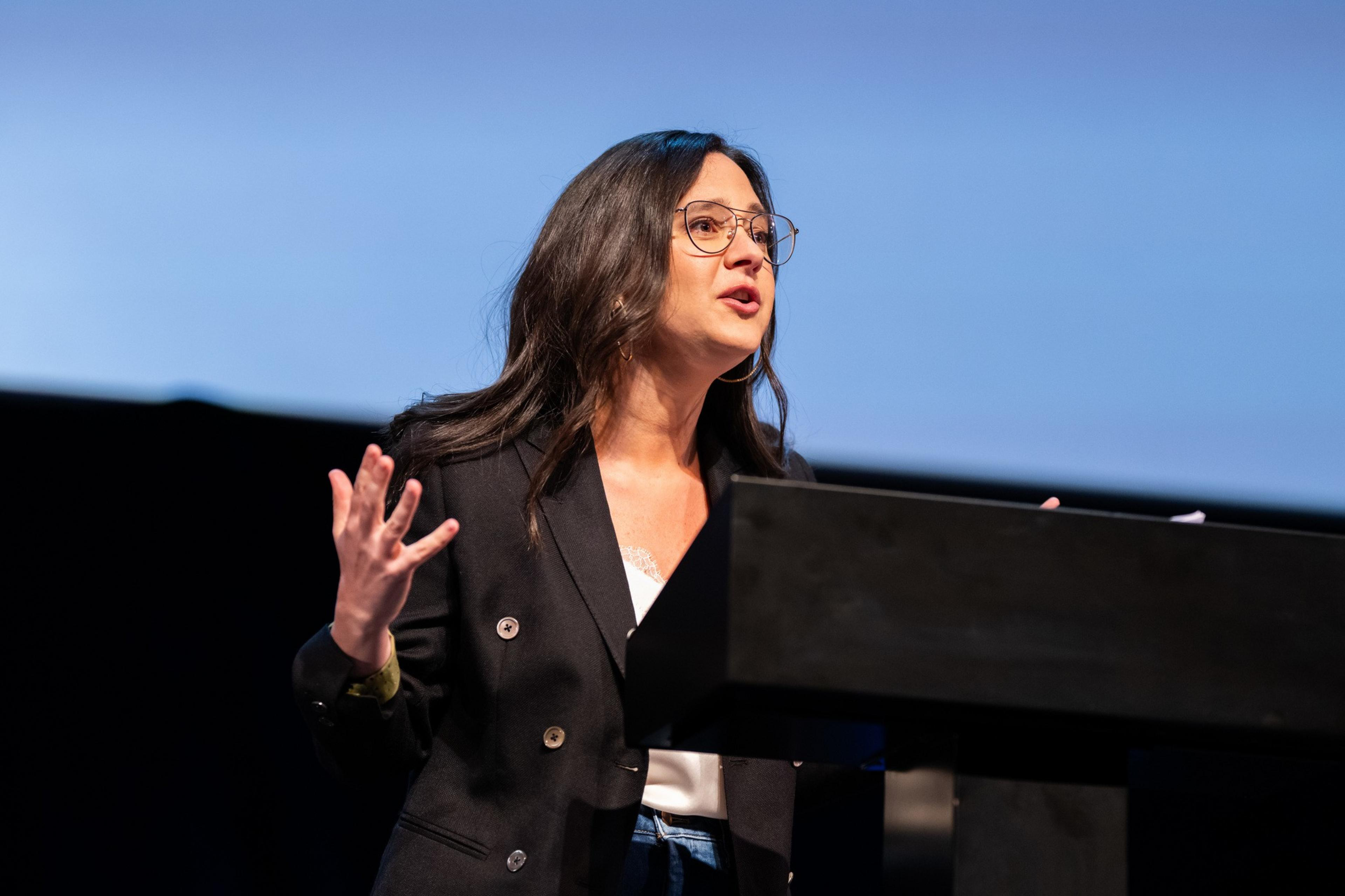 A woman with long dark hair and glasses is speaking passionately at a podium, wearing a black blazer over a white top, with a blue background behind her.
