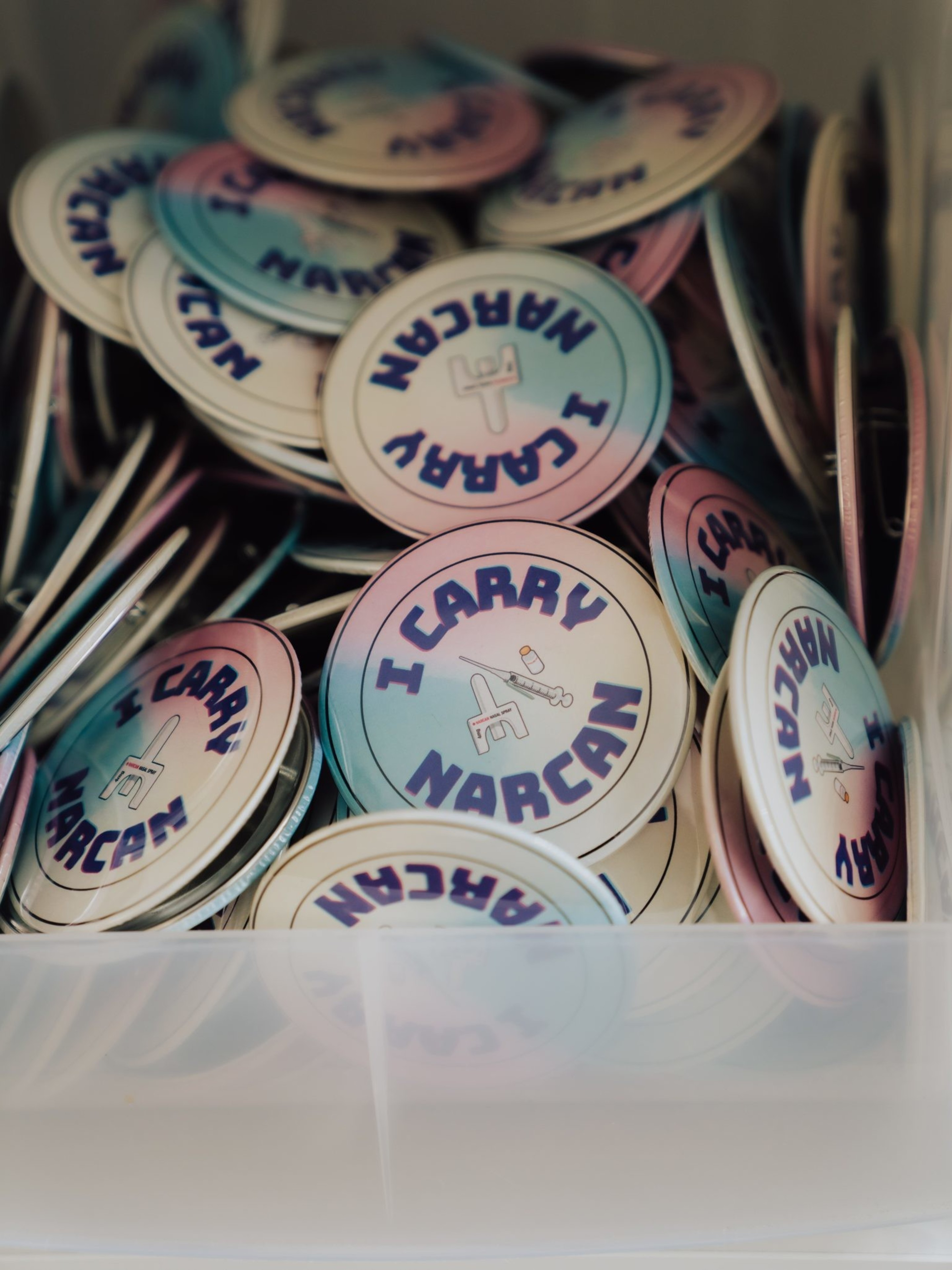 The image shows a container filled with round buttons that say &quot;I CARRY NARCAN,&quot; featuring a syringe graphic in the center with a pink-to-blue gradient.