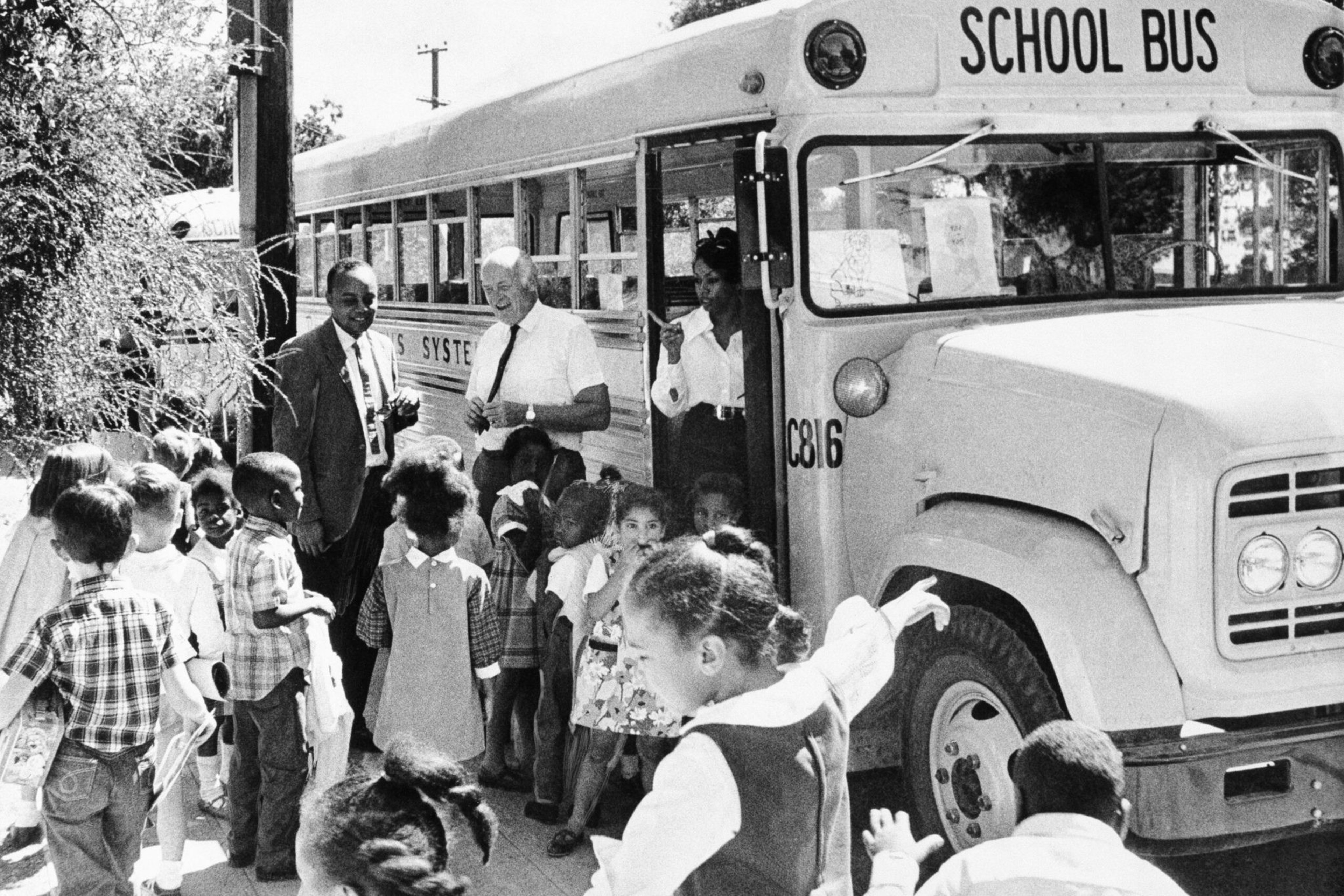 Children of diverse backgrounds wait and line up to board a school bus, while two men and a woman stand nearby assisting.