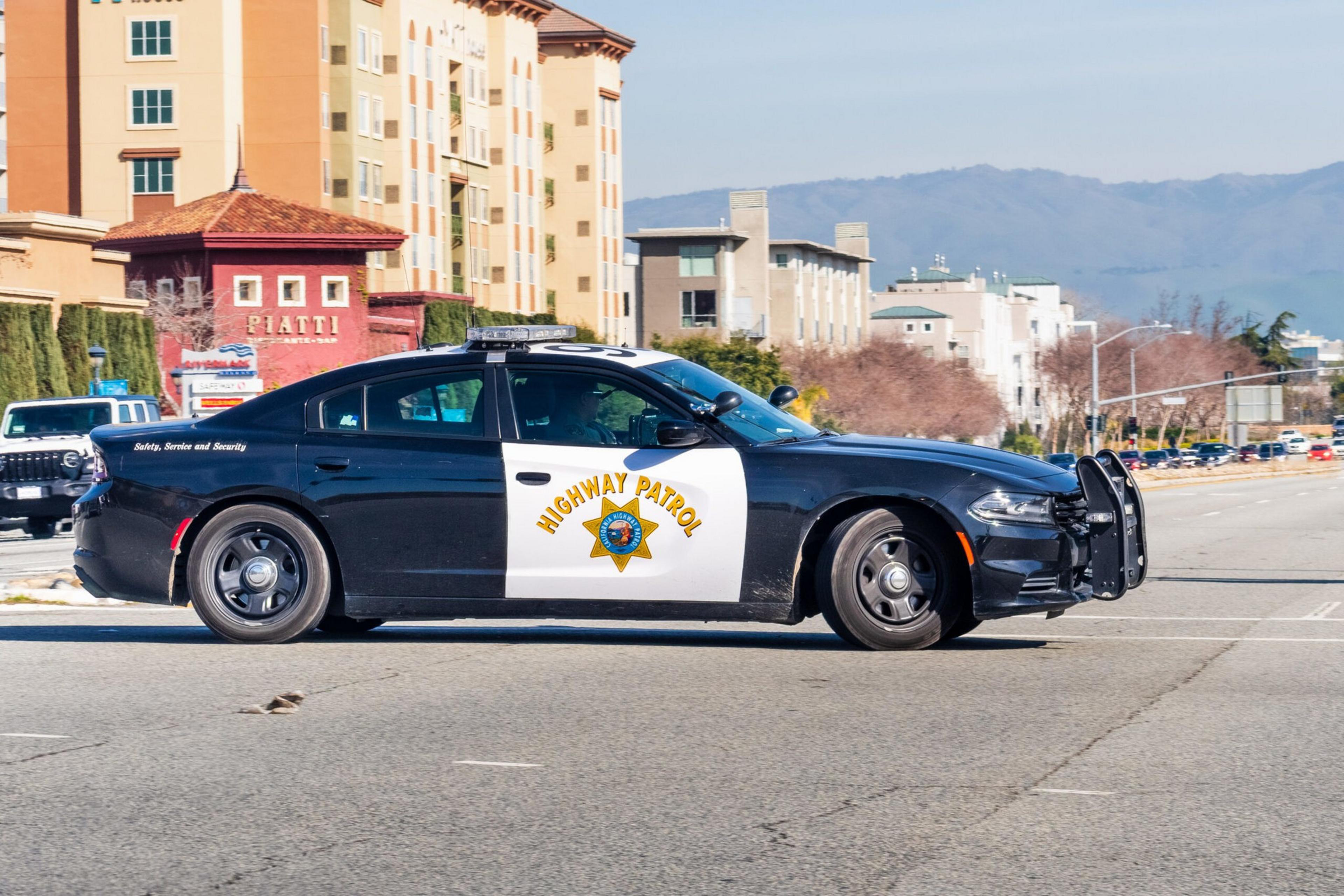 A black and white highway patrol car is parked on a city street. The background features a few buildings, trees, and distant hills.