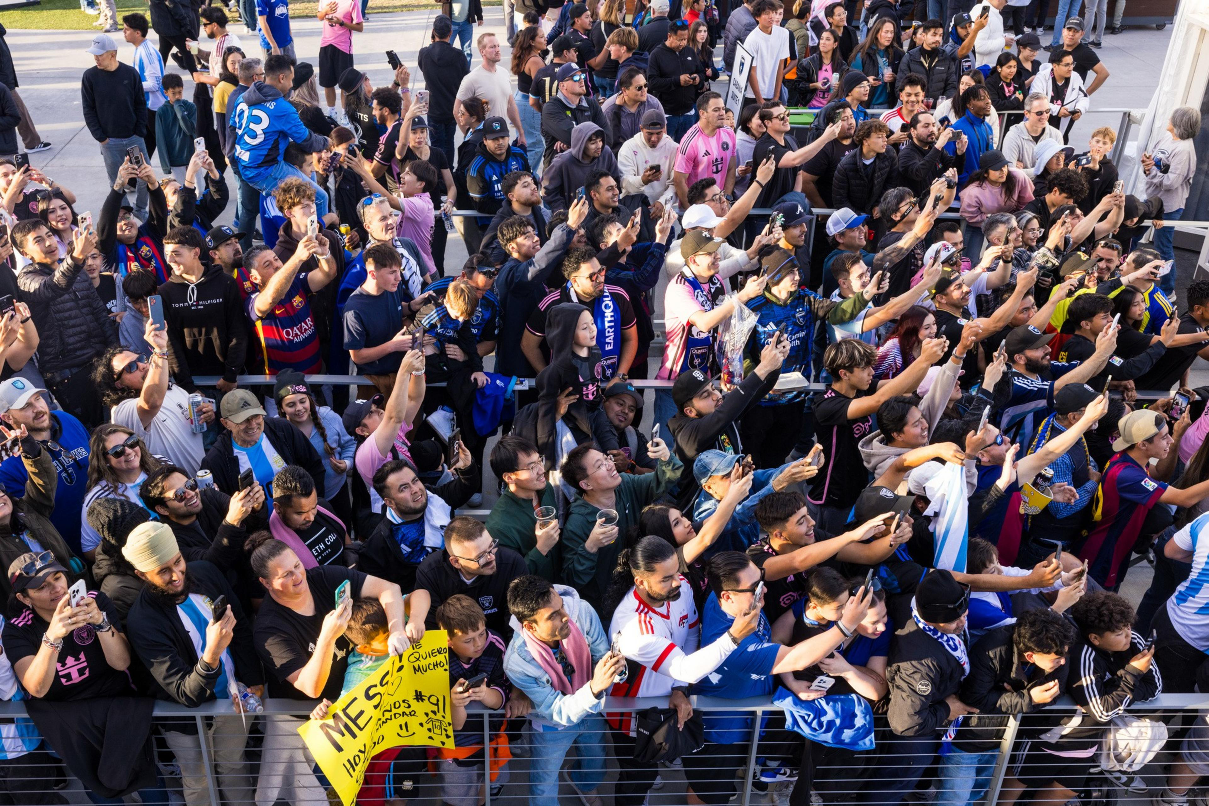 A large crowd of enthusiastic fans, mostly in sports jerseys, eagerly take photos and videos, some holding signs and drinks, creating a lively and excited atmosphere.