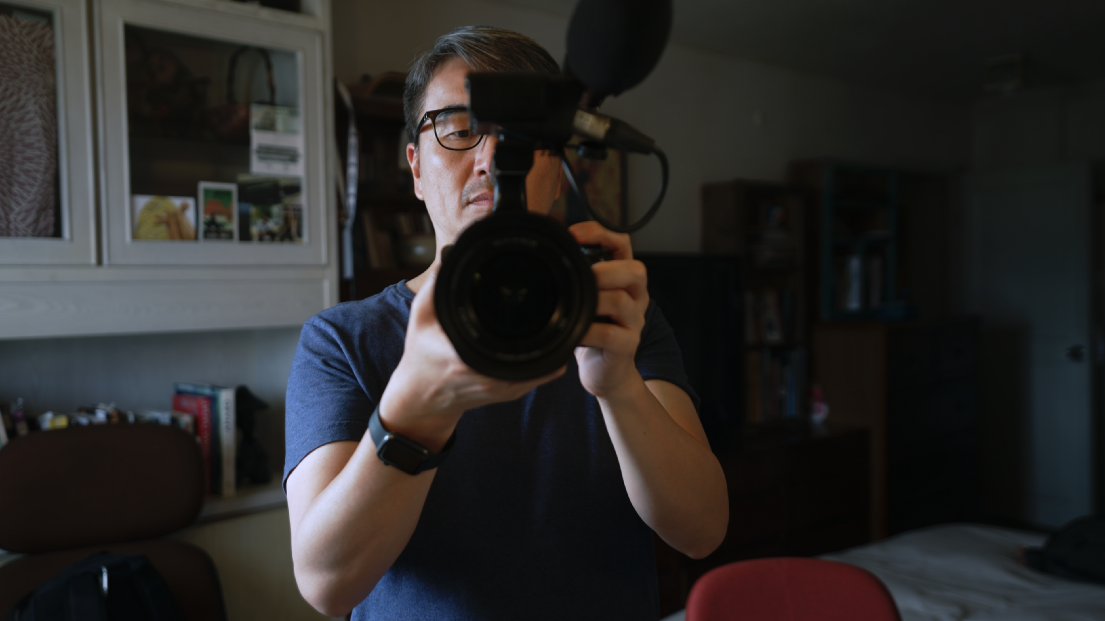 A man holding a camera with a microphone attached, aiming it forward in a room with bookshelves.
