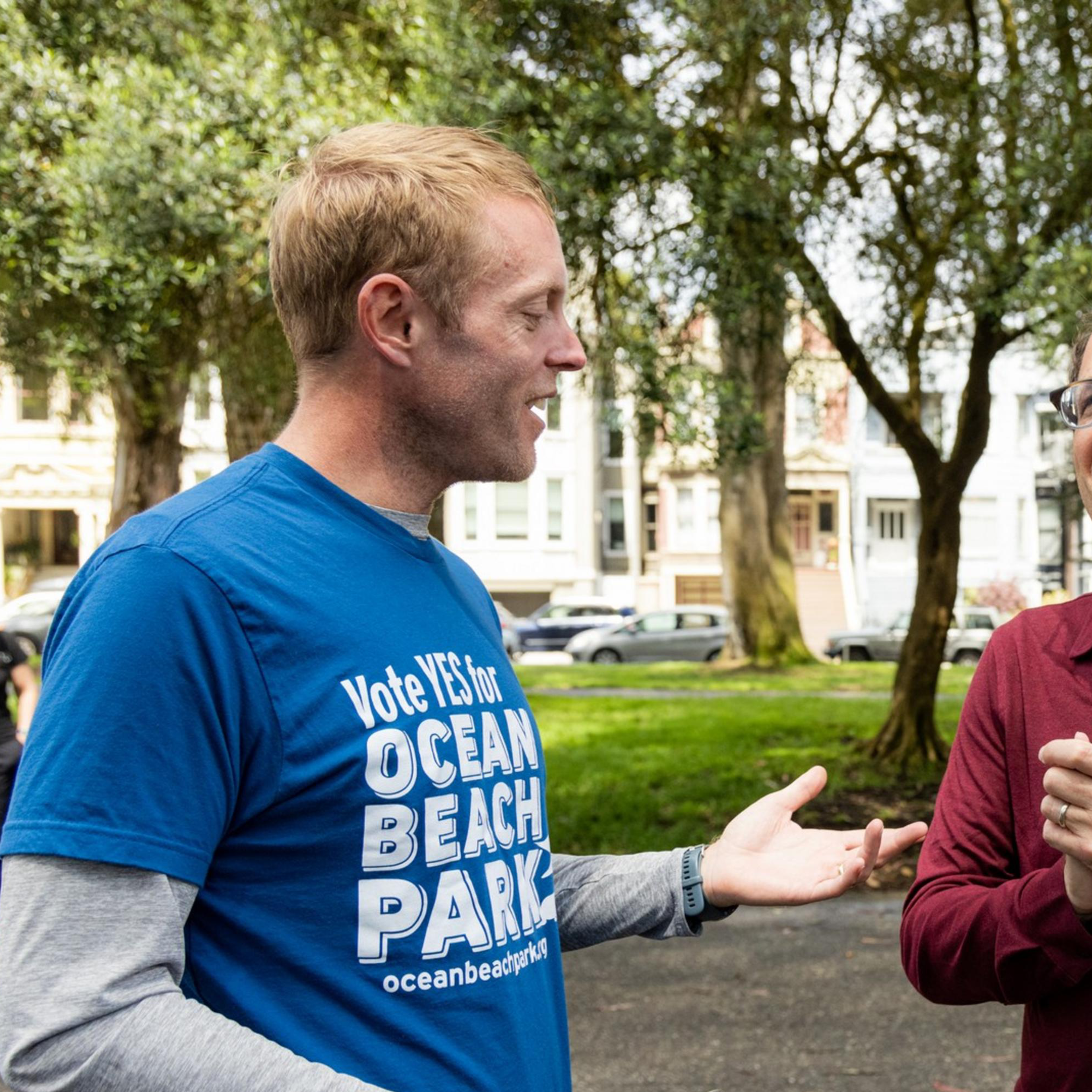 Two men conversing in a park, one in a blue &quot;Vote YES for Ocean Beach Park&quot; shirt, the other in a red shirt, while another man in a similar blue shirt walks behind them.