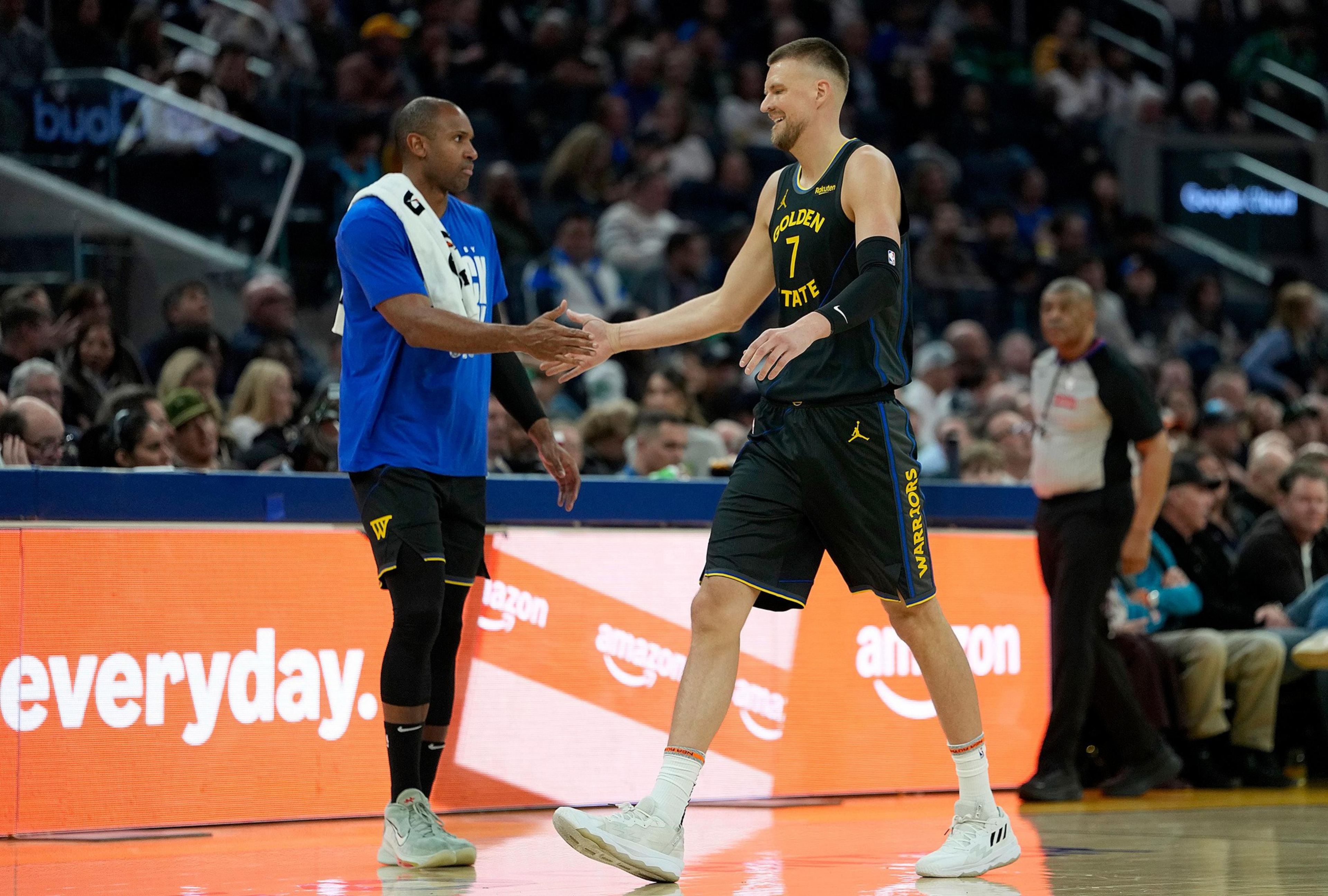 Two basketball players from the Golden State Warriors shake hands on the court during a game, with a crowd and referee in the background.