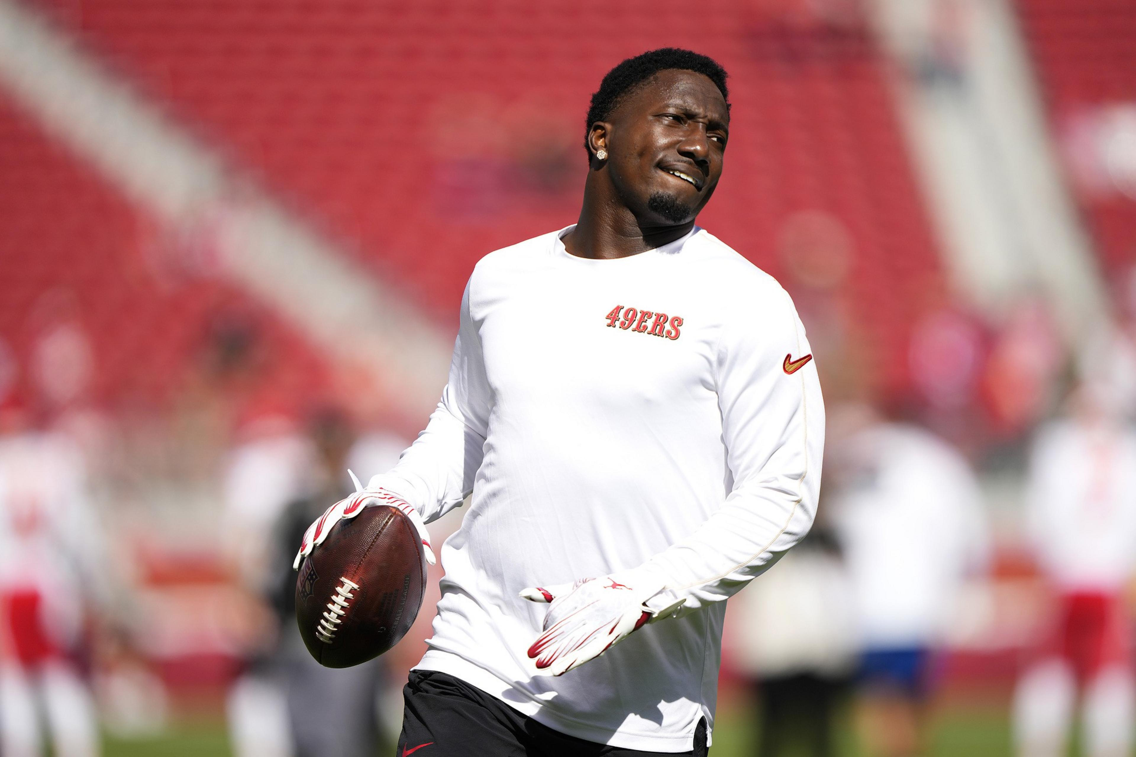 A football player in a white 49ers shirt holds a football on a field. He looks sideways with a slight smile. The background is a blurred stadium.