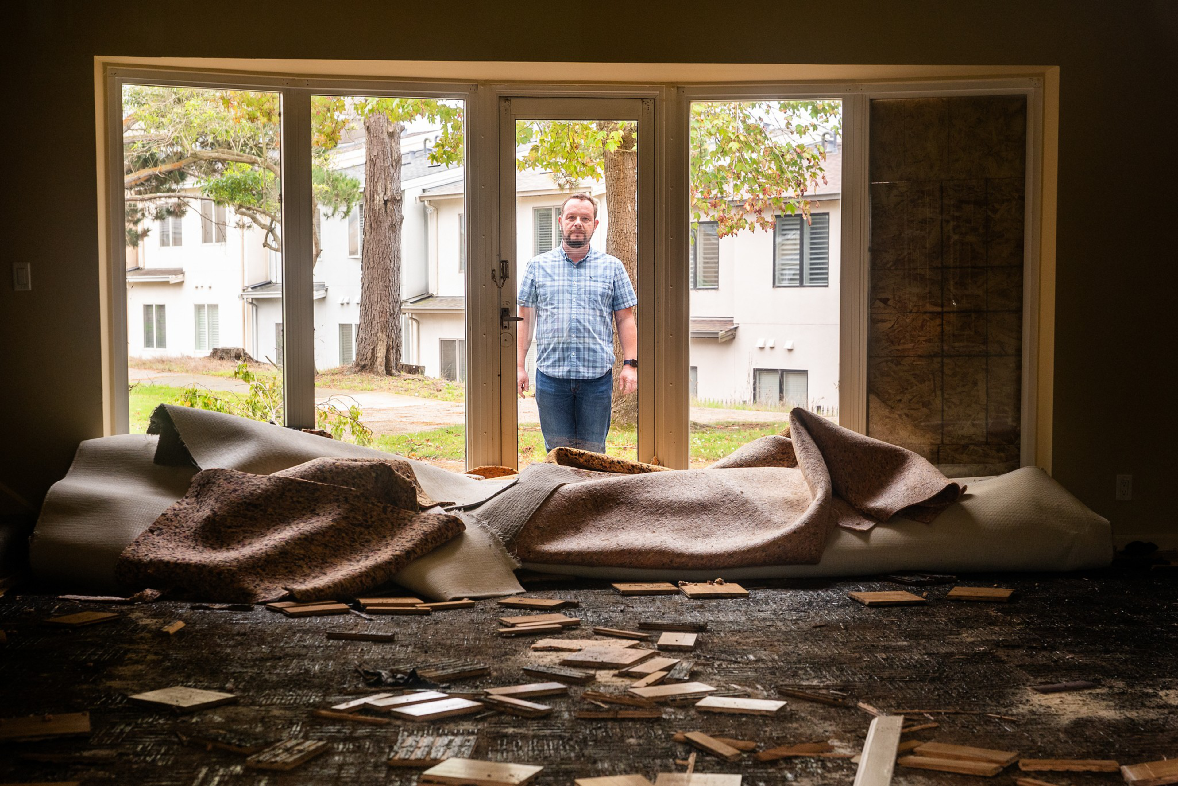 A man in a blue plaid shirt stands outside a large window looking into a room with torn-up carpet and scattered wooden floor pieces.