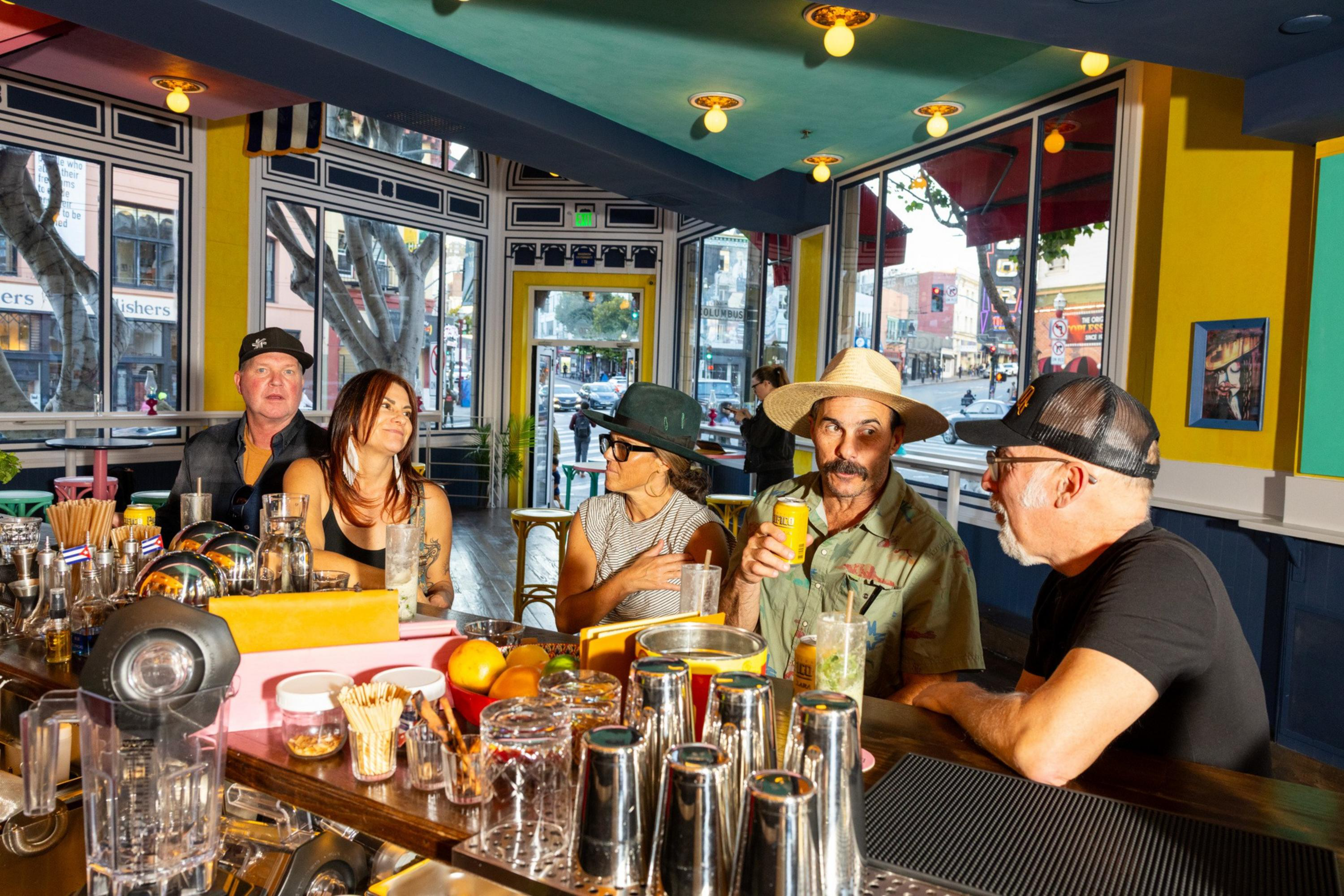 Five people sit at a brightly lit bar with drinks, engaged in conversation, with large windows showing a street scene outside.