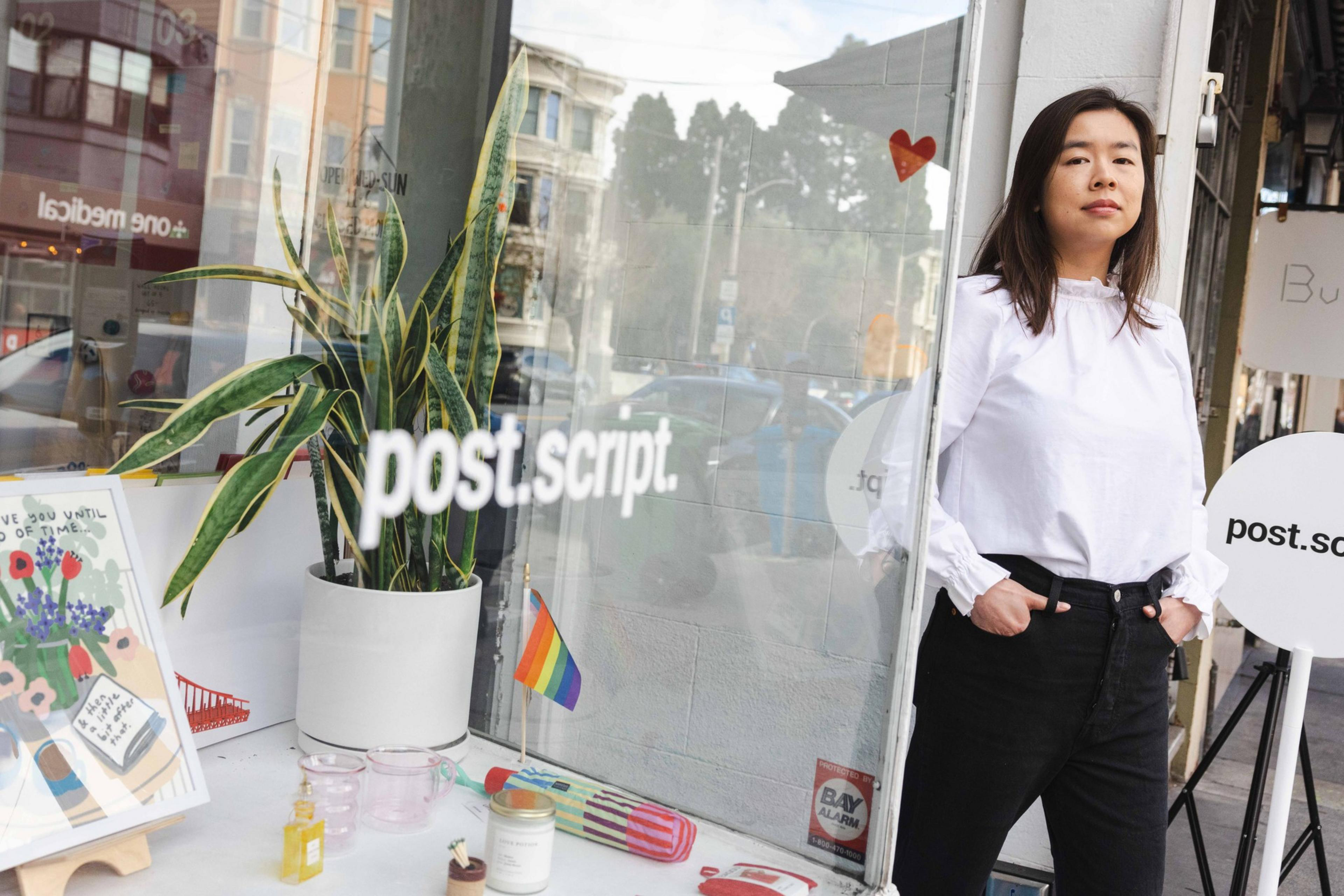 A woman in a white shirt stands outside a shop named "post.script." The window displays a plant, small items, a poster, and a rainbow flag.