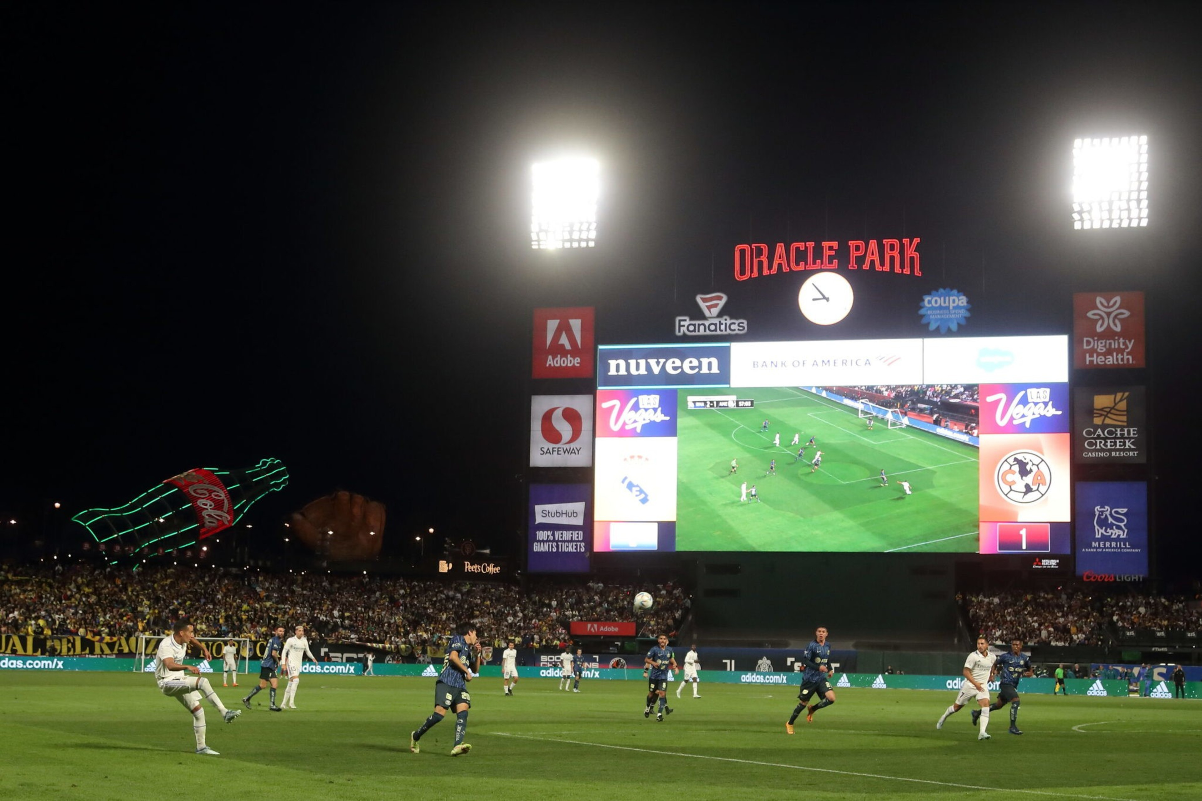 A night soccer match at Oracle Park with players on the field and a large scoreboard displaying the game. Bright lights and a Coca-Cola bottle add to the scene.