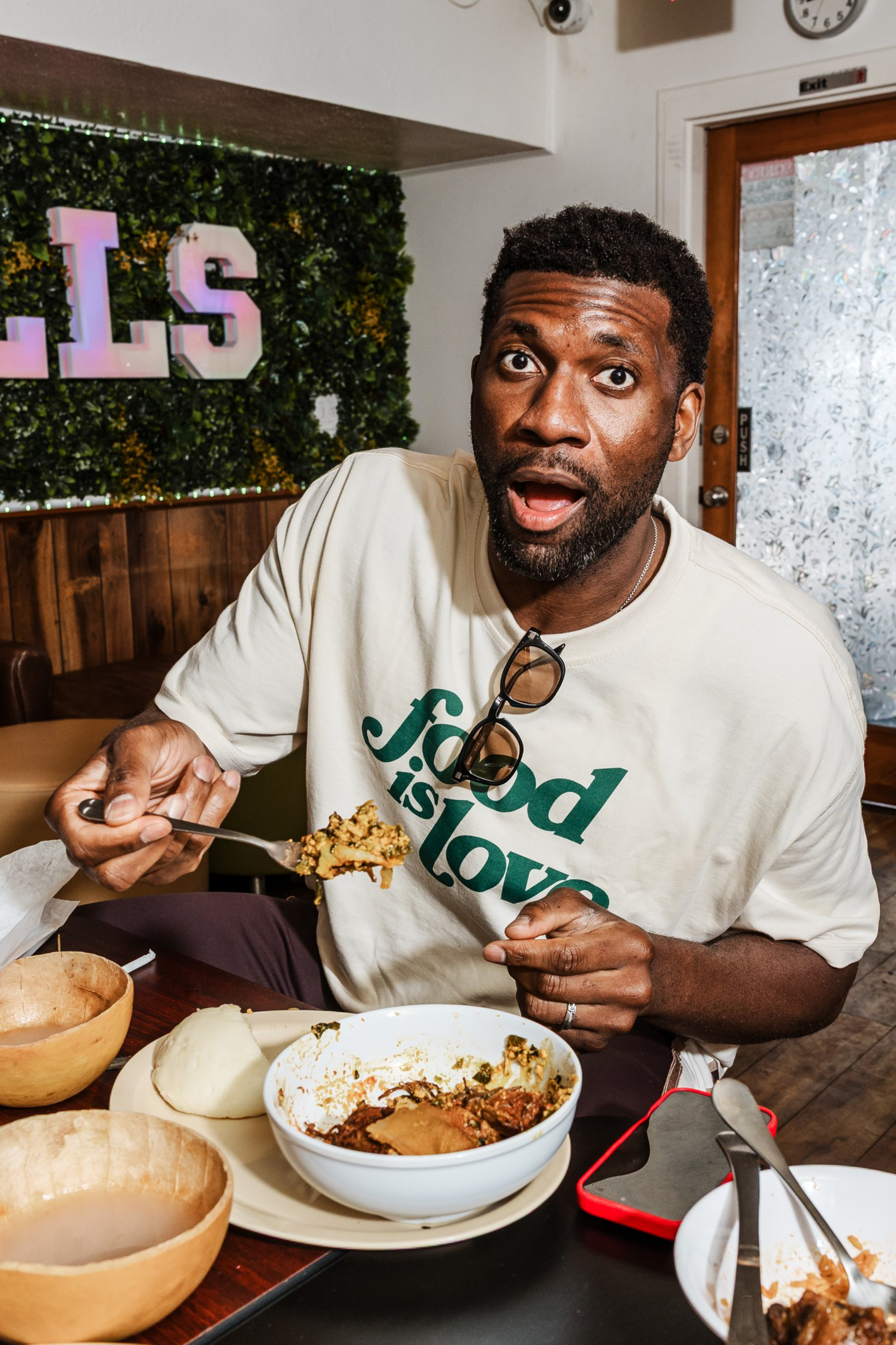 A man wearing a “food is love” shirt looks surprised while holding a spoonful of food over a bowl at a table with dishes and a red phone.