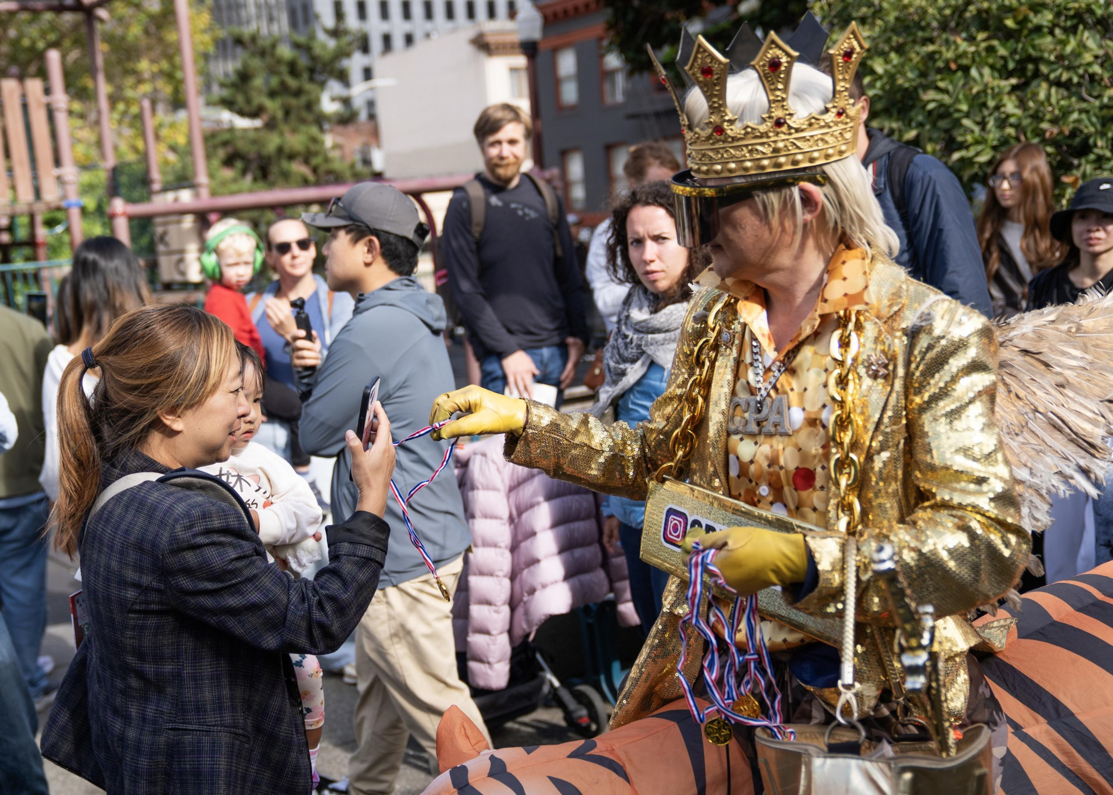 A person dressed in a gold crown, gold outfit, and wings hands medals to a woman holding a phone, with multiple people watching in the background.