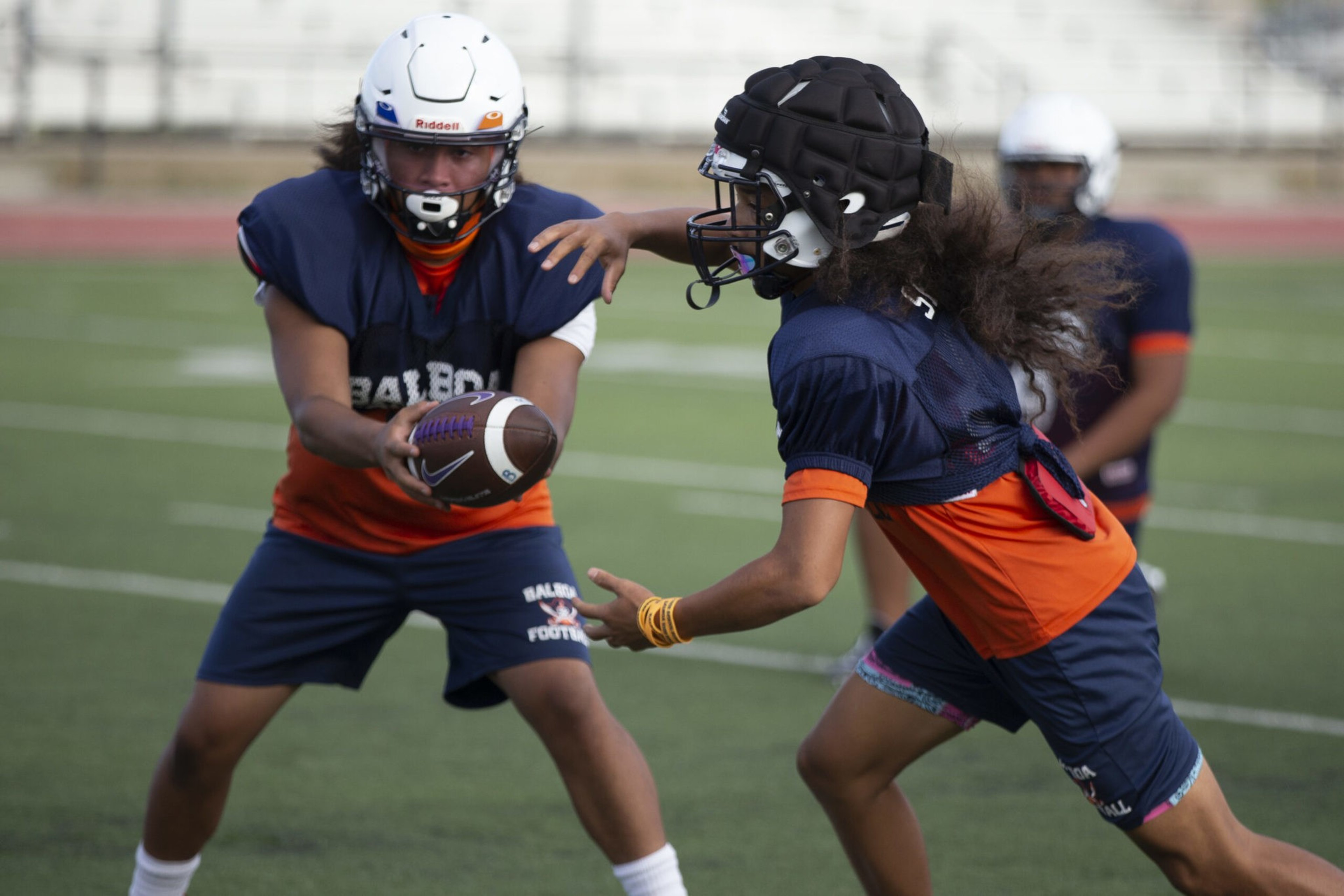Two young football players in helmets and uniforms pass a football on a field, with one player reaching to catch it and another running nearby.