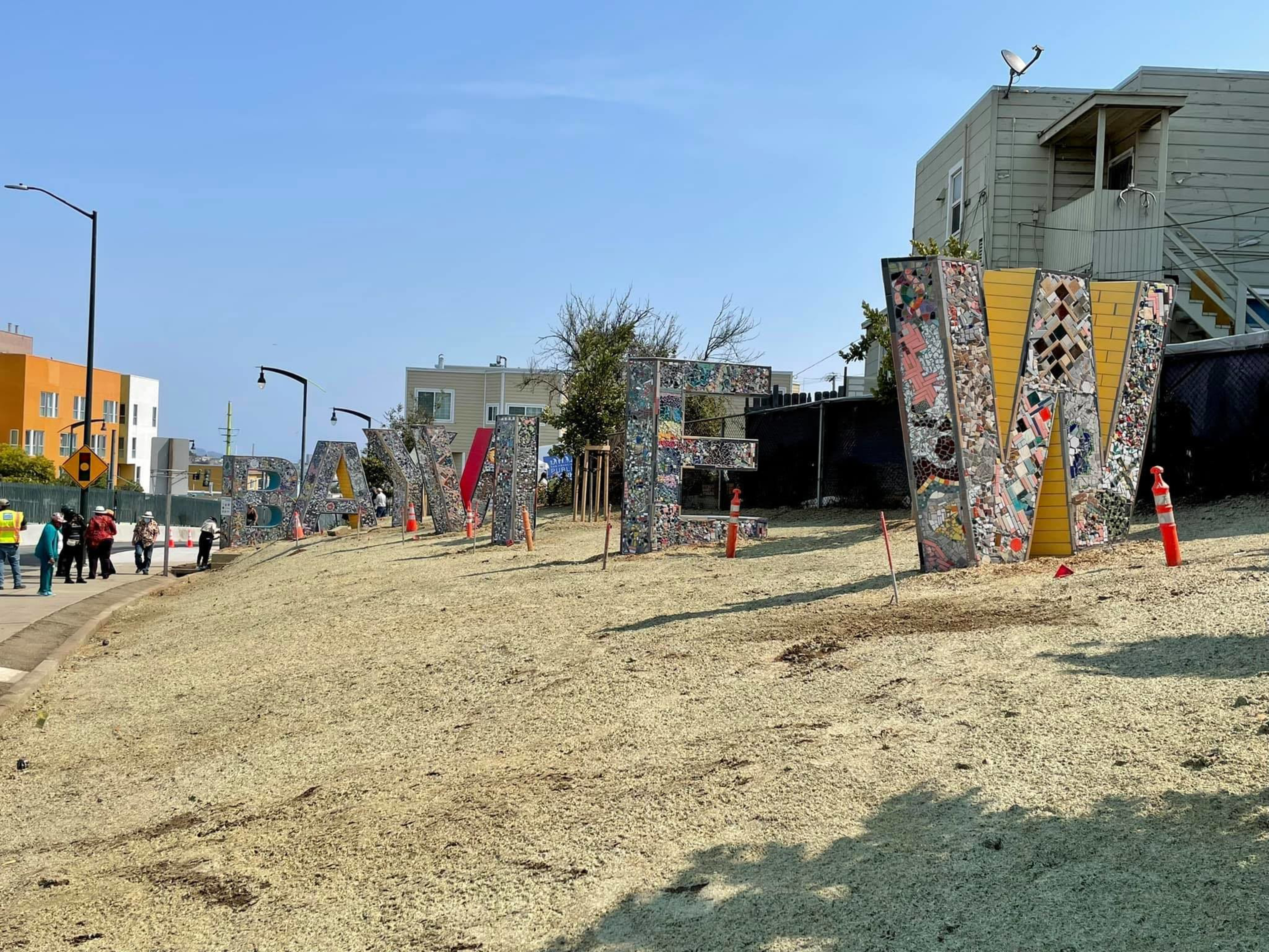 Large mosaic-covered letters spelling "BAY VIEW" stand on a grassy hill beside a sidewalk with people walking nearby under a clear blue sky.
