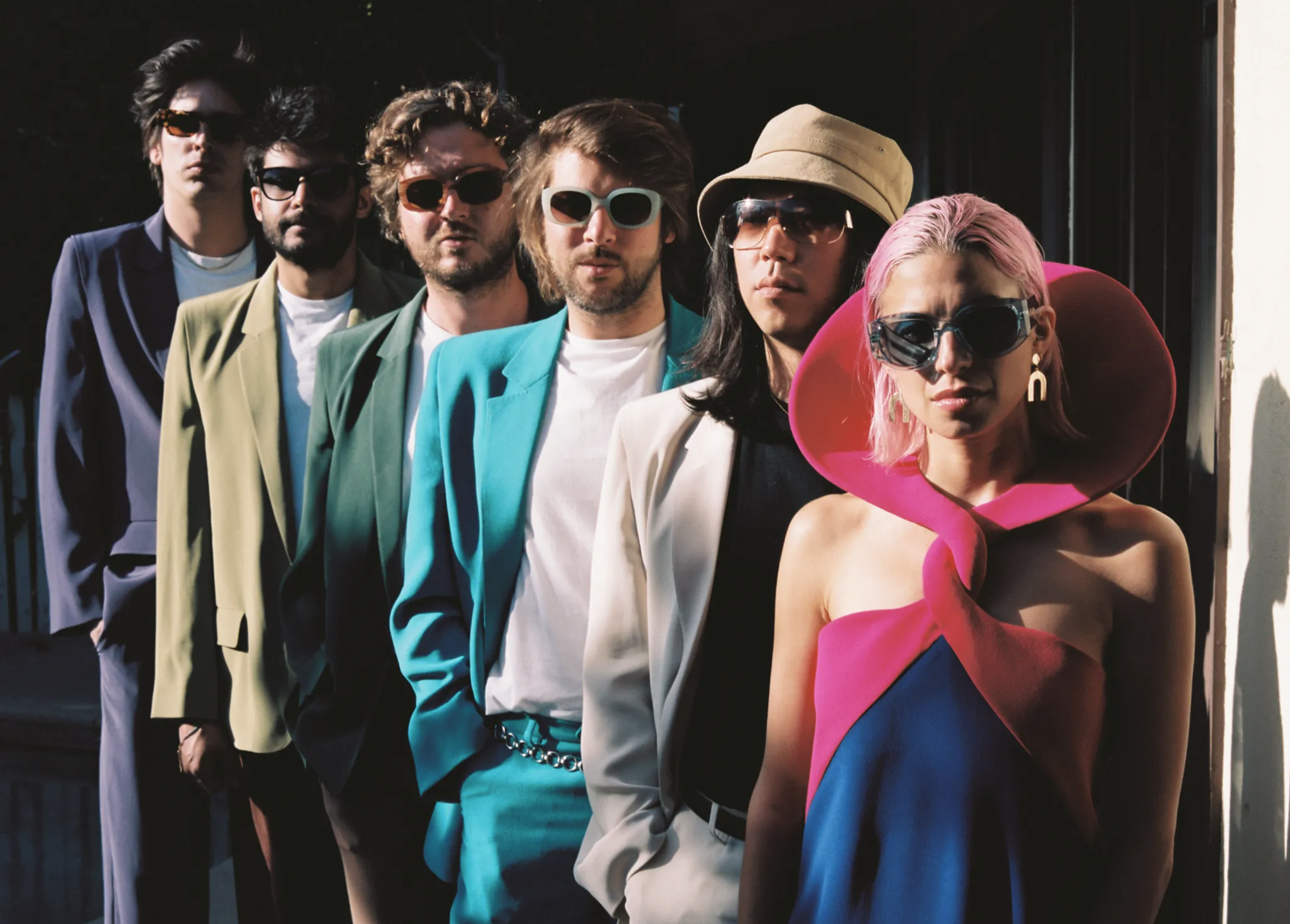 Six people stand in a line, each wearing sunglasses and colorful, stylish outfits, with the person in front sporting pink hair and a unique collar.