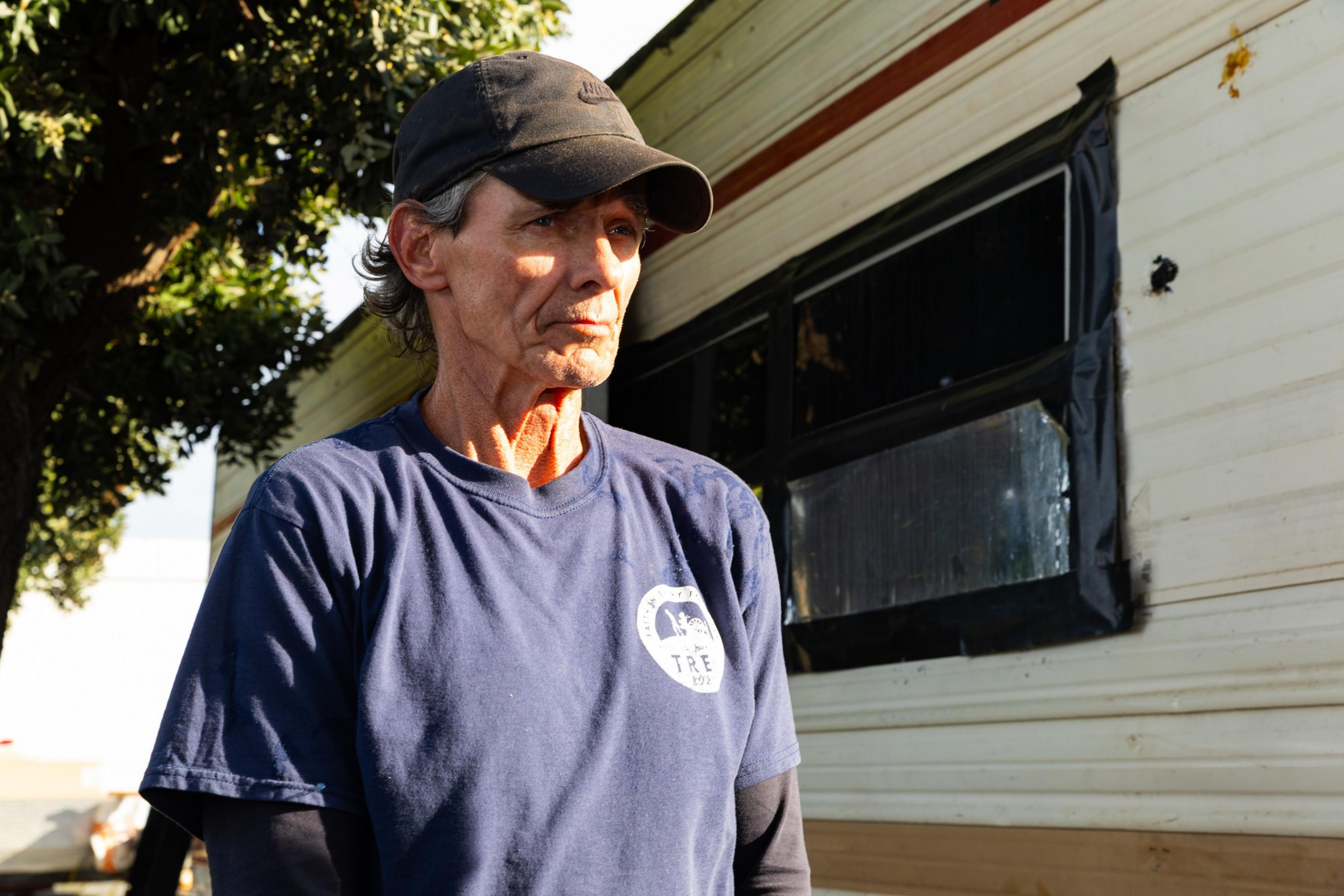 A person wearing a blue shirt and black cap stands outside near a tree and an RV with taped windows, illuminated by sunlight.