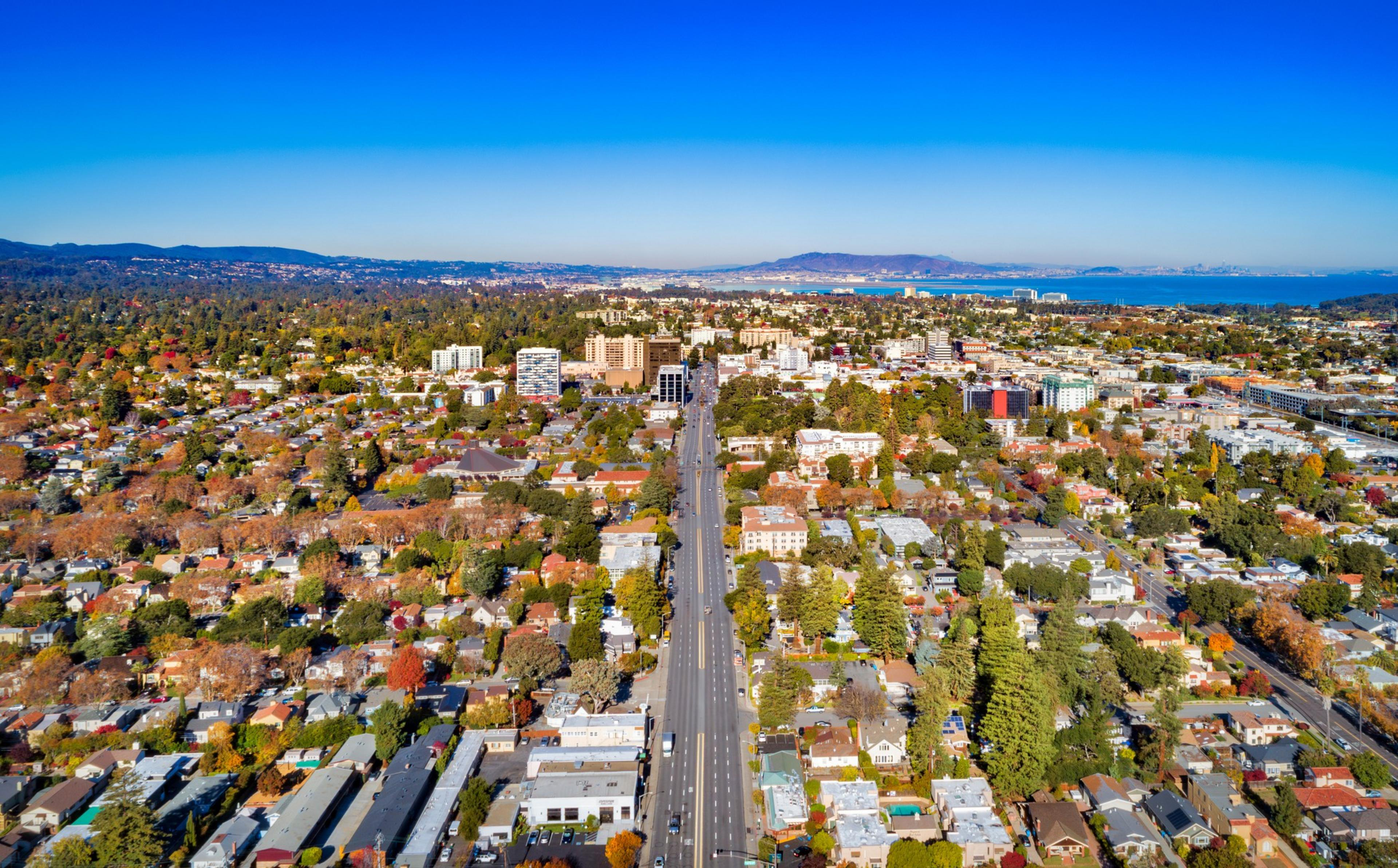 An aerial view of a city shows a straight road leading into a dense area with buildings, surrounded by green trees, with mountains and a body of water in the background.