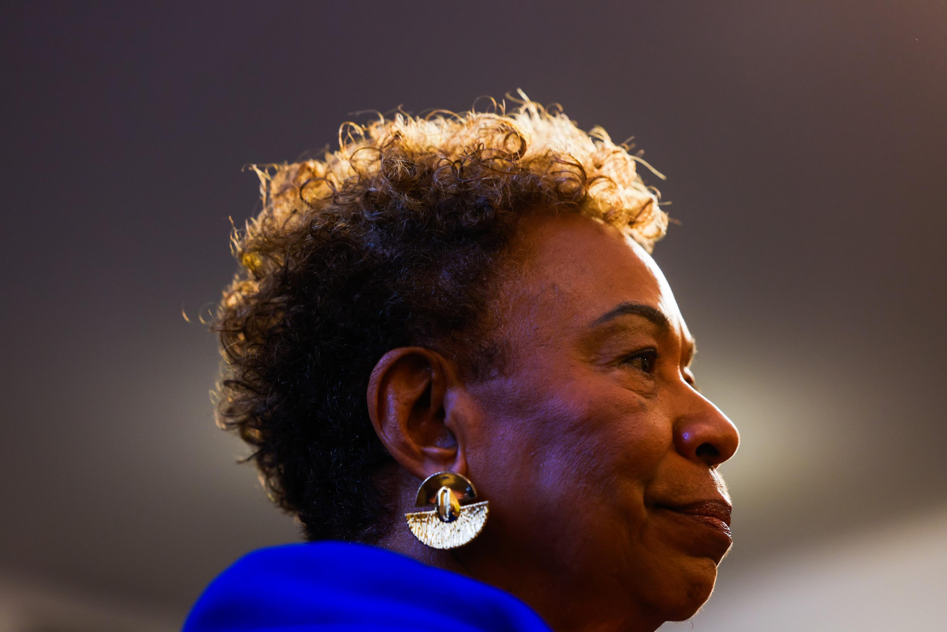 A woman with short curly hair and large, gold, fan-shaped earrings faces right, wearing a bright blue garment against a soft, blurred background.