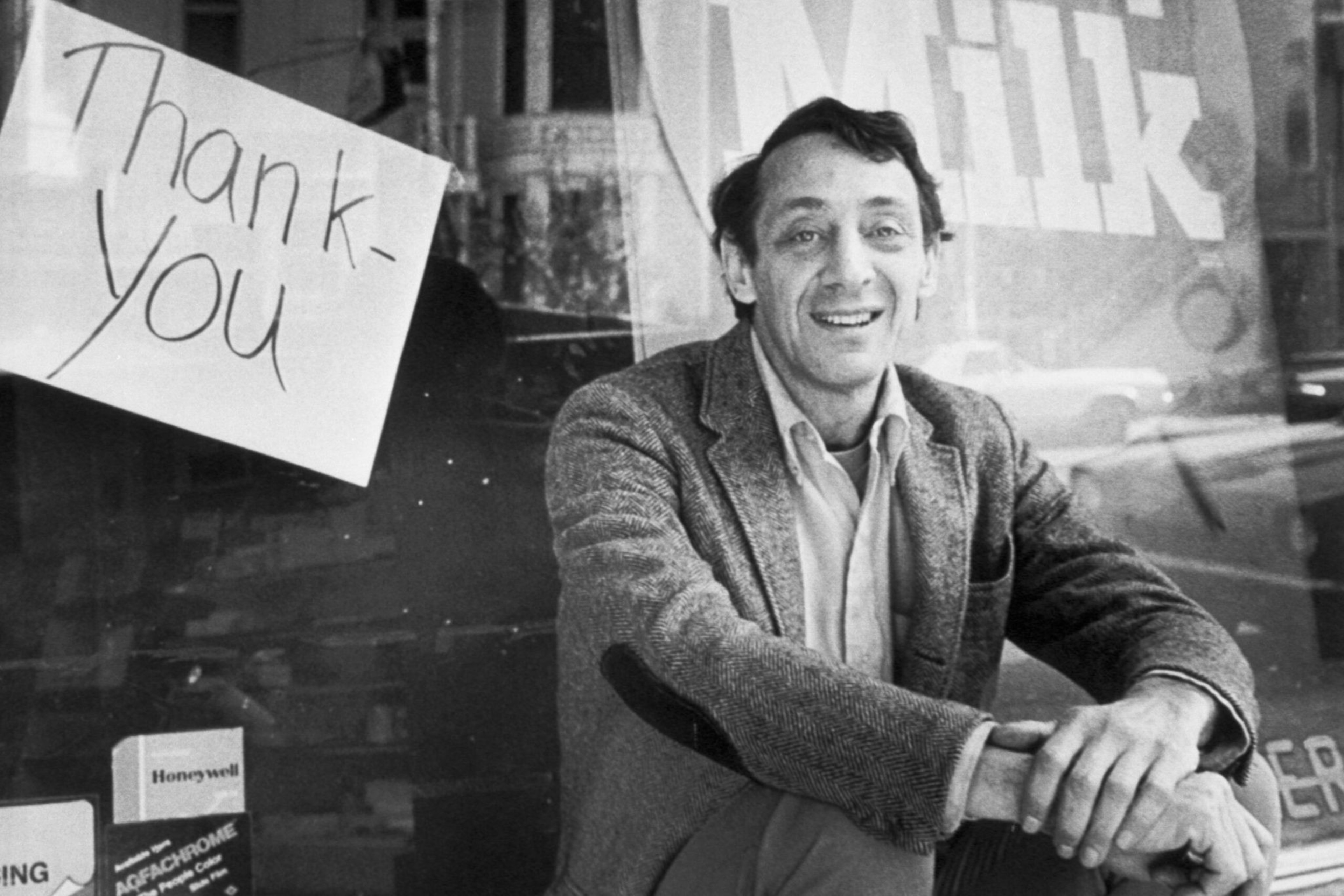 A man in a blazer smiles while sitting near a window with a "Thank you" sign taped on it.