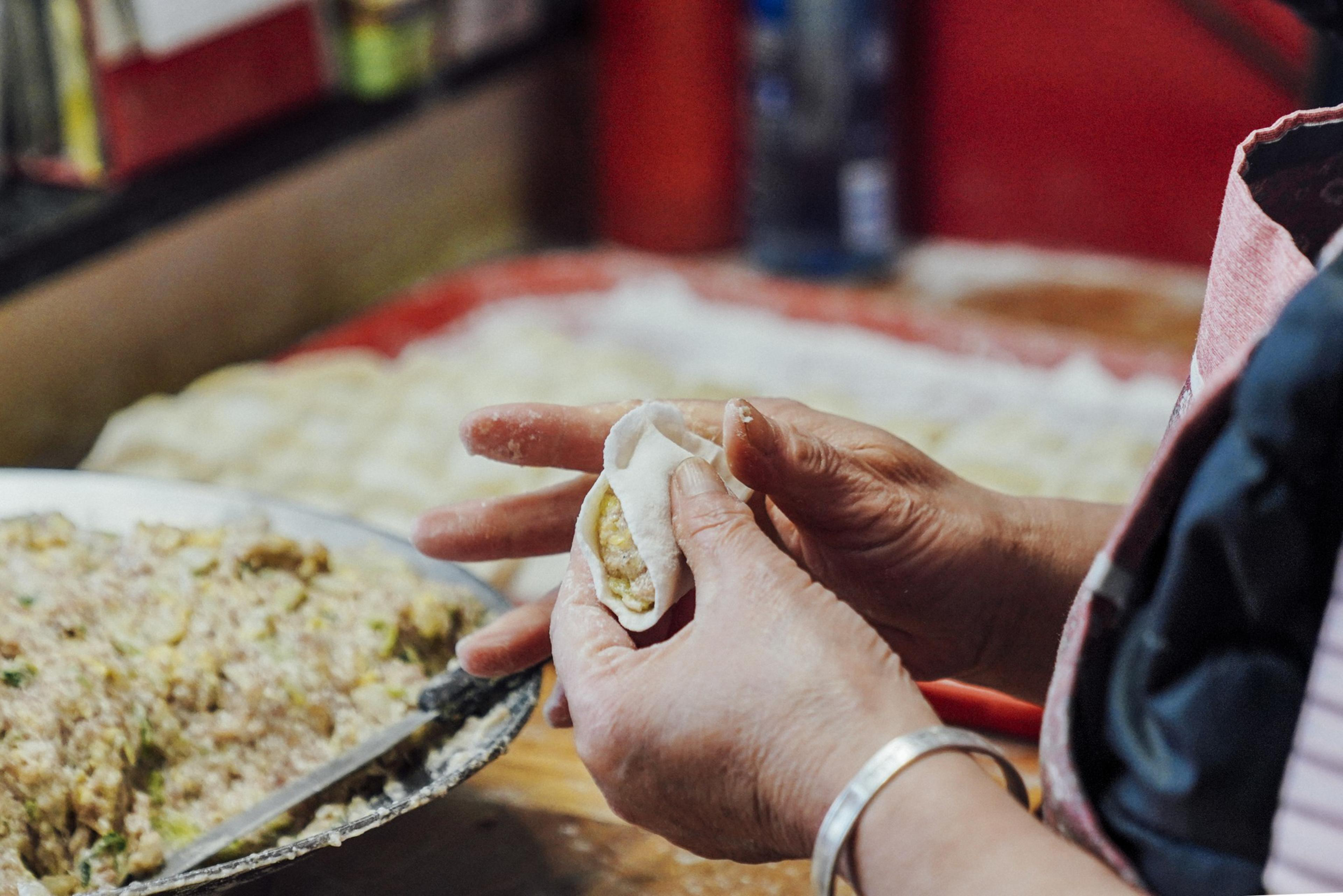 A person's hands are shaping dumplings. They're holding a dumpling wrapper with filling inside, and a plate of filling is nearby.