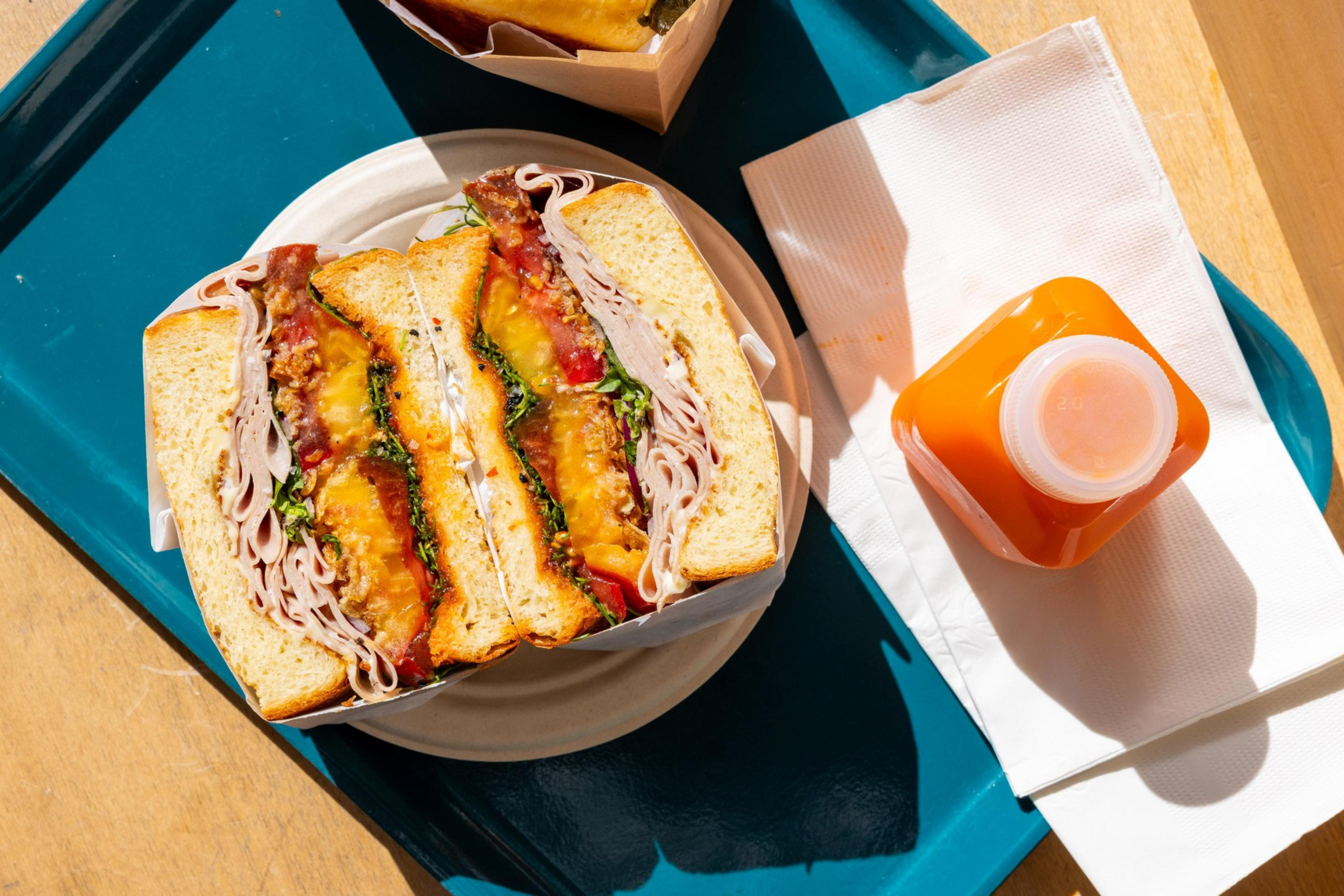 A turkey sandwich with cheddar, lettuce, tomato, and bacon on white bread sits on a plate with a small bottle of orange juice and a napkin on a blue tray.