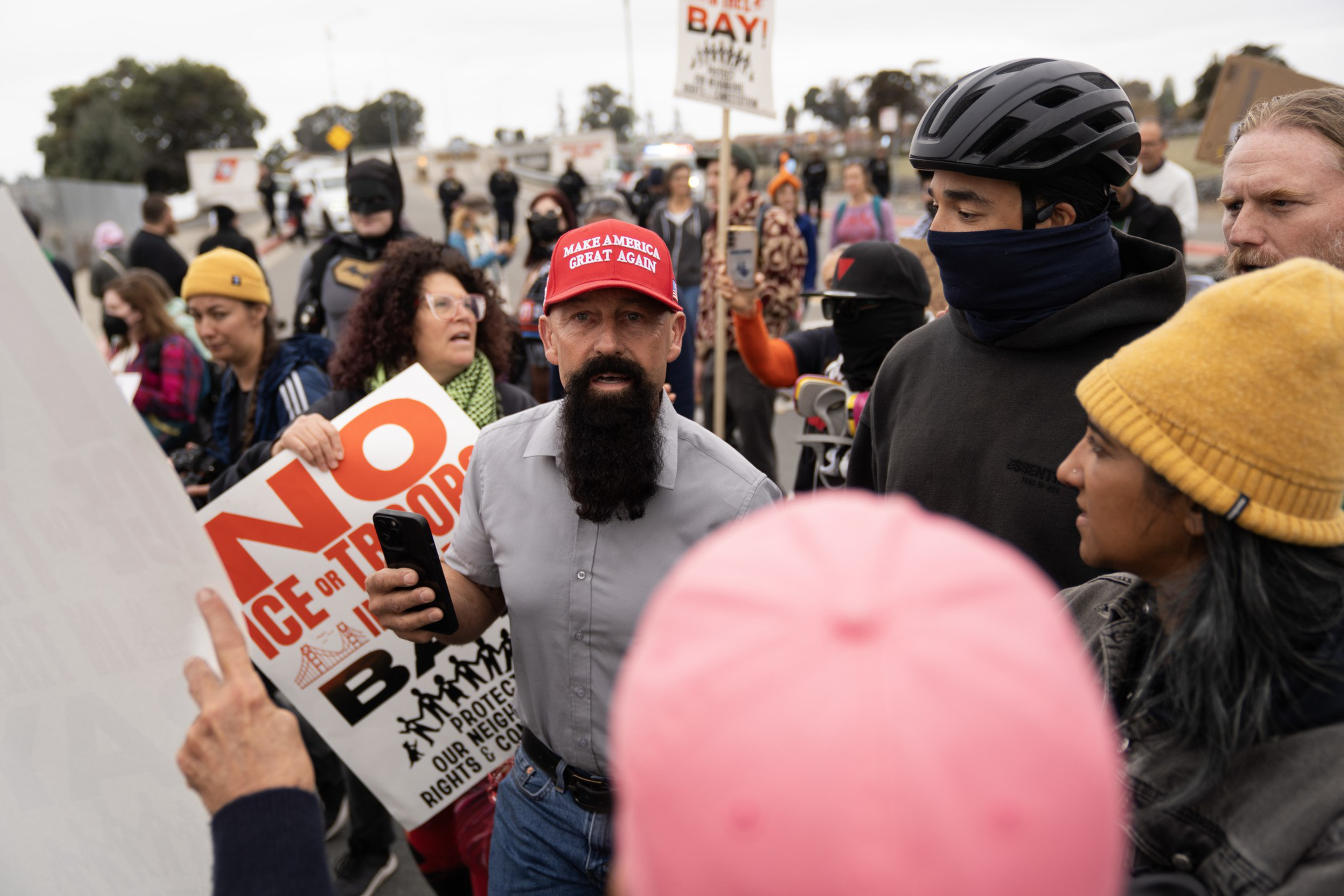 A bearded man in a red “Make America Great Again” hat faces a crowd holding protest signs, with people wearing hats and face coverings nearby.